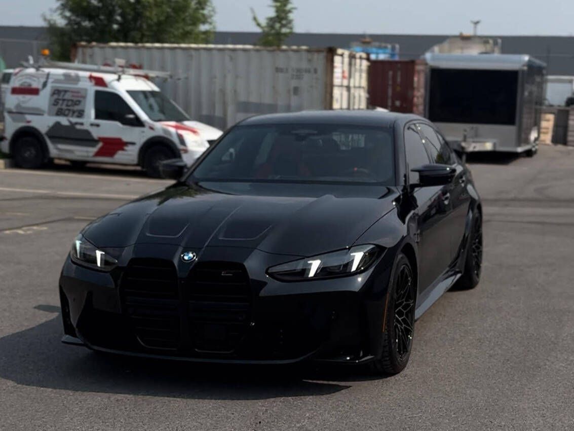 Black BMW sedan parked in a lot, with an ambulance and industrial trailer in the background