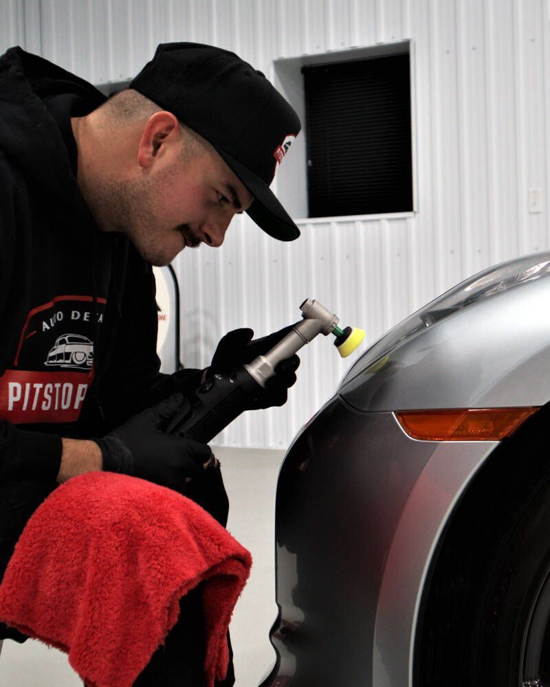 Detailer polishing a silver car's front fender with a buffer, red towel in hand, in a workshop