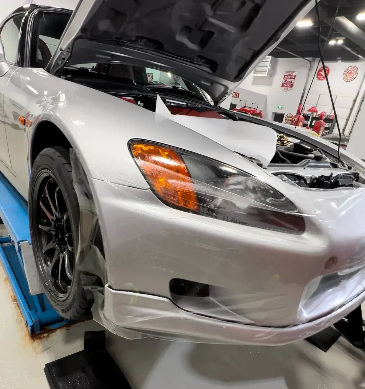 Silver sports car with hood open in a workshop, showing front damage and black wheels on a lift.