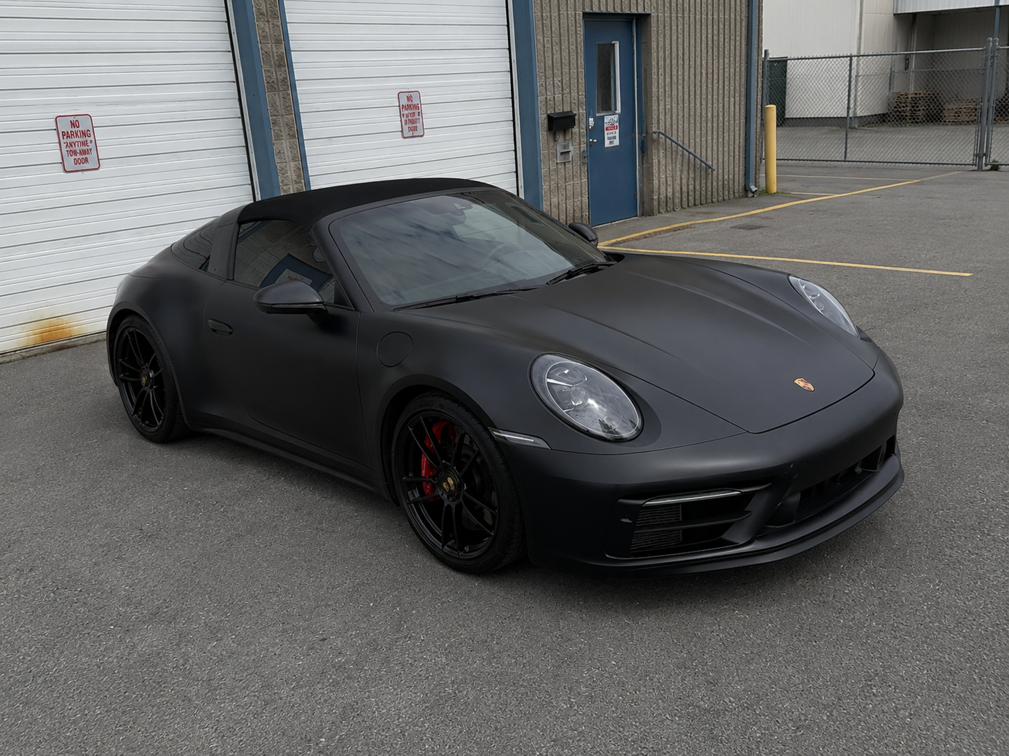 Black Porsche sports car parked outside an industrial building, matte finish with red brake calipers.