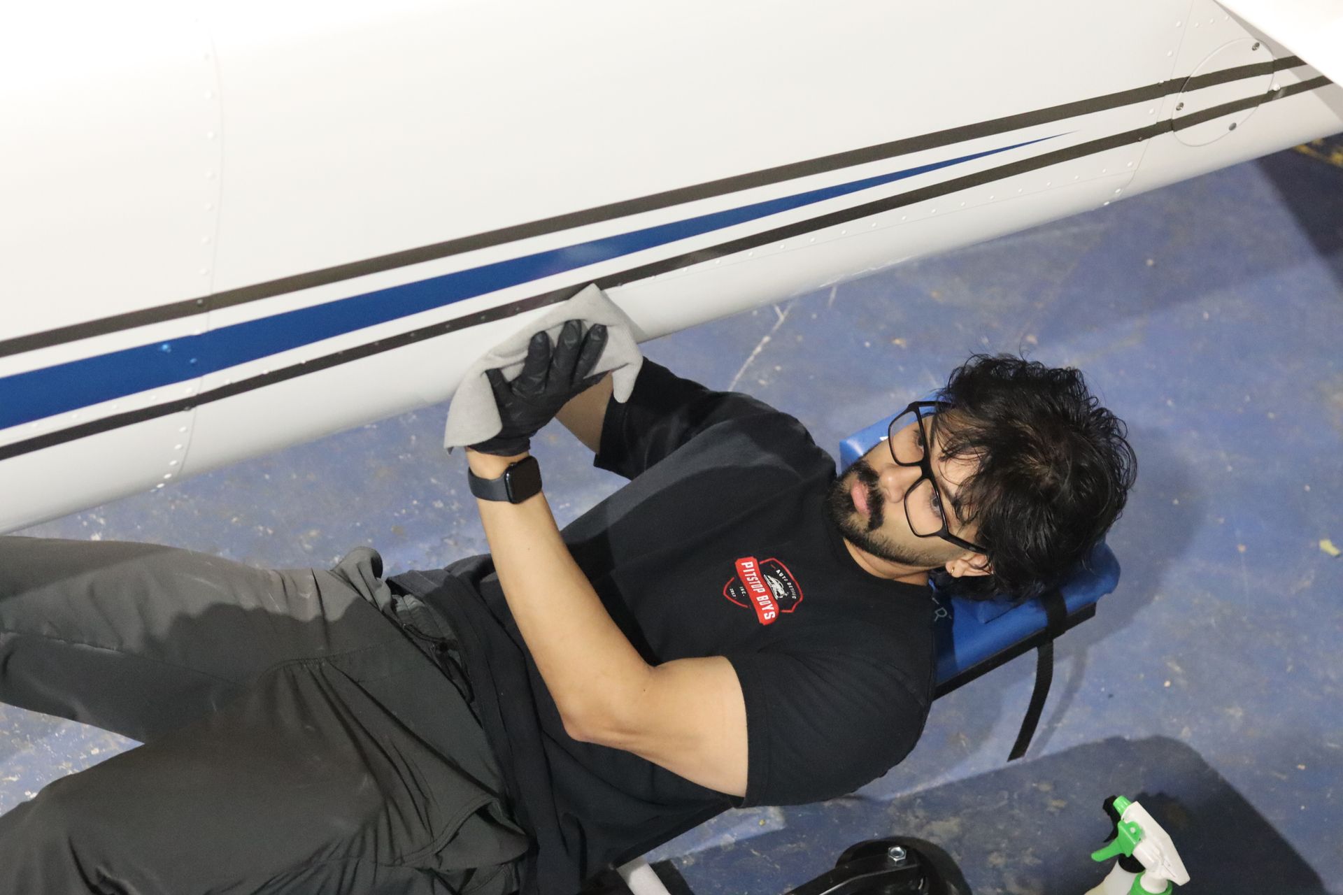 Two people working under a small airplane in a hangar, with tools and a white bucket nearby.