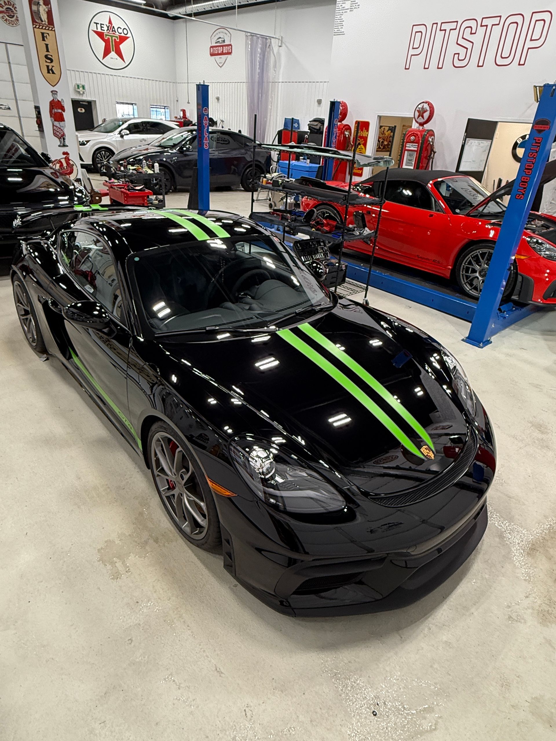 Black sports car with green racing stripes in an auto workshop, with a red car on a lift in the background
