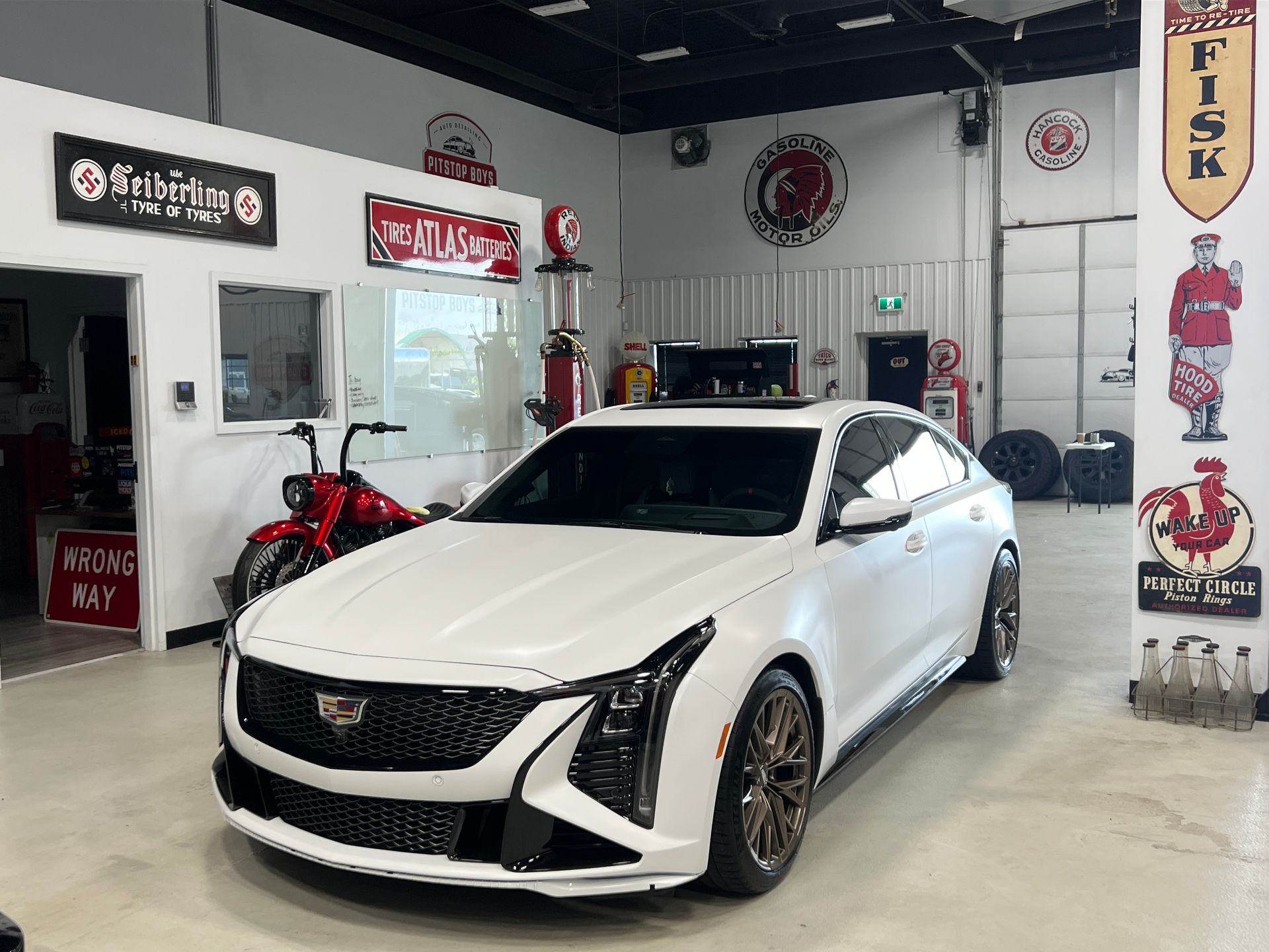 White Cadillac coupe in a garage showroom, surrounded by auto shop signs and equipment.