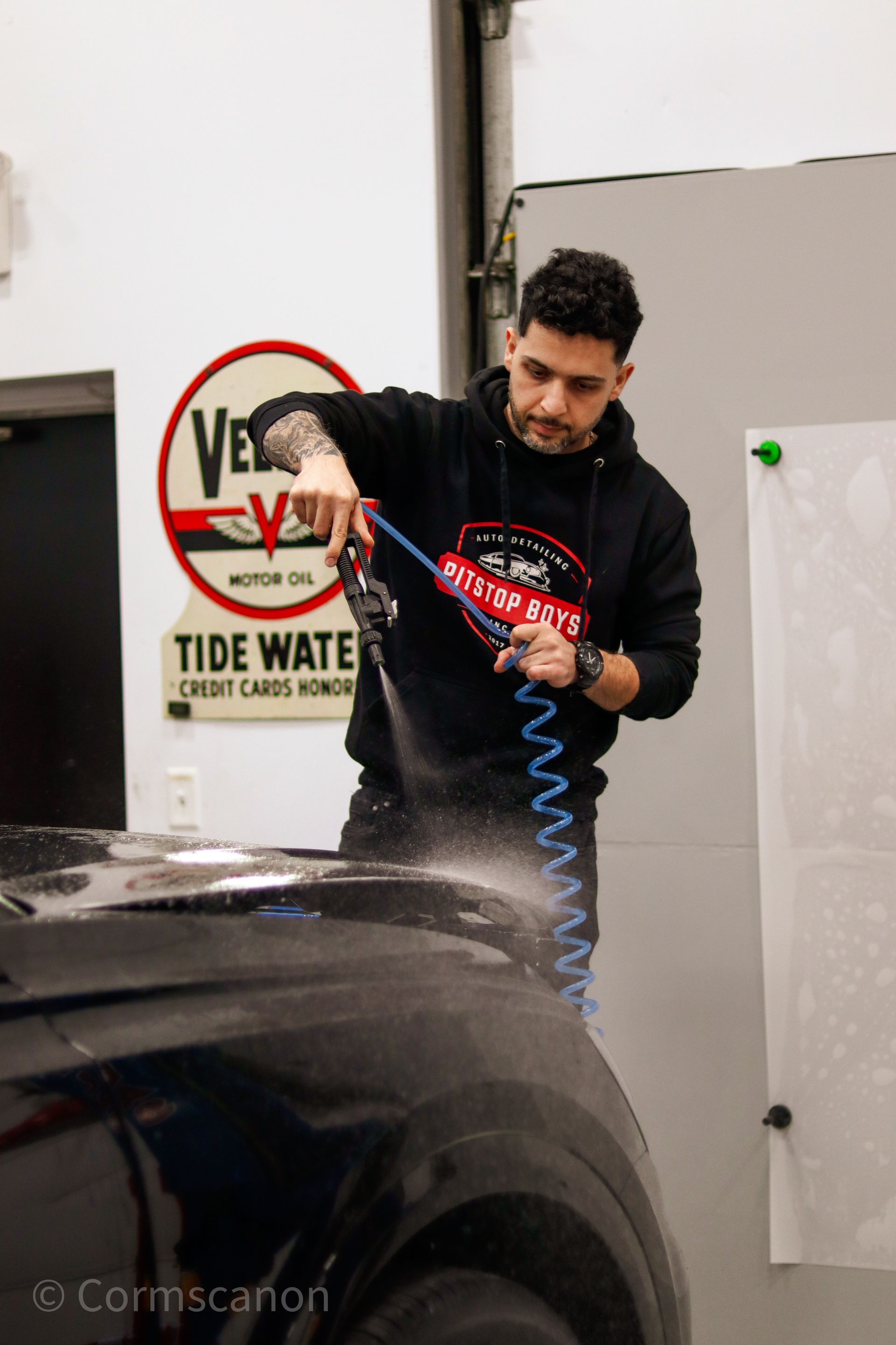 Man in a black hoodie polishing a car with a red sponge indoors near a wall sign