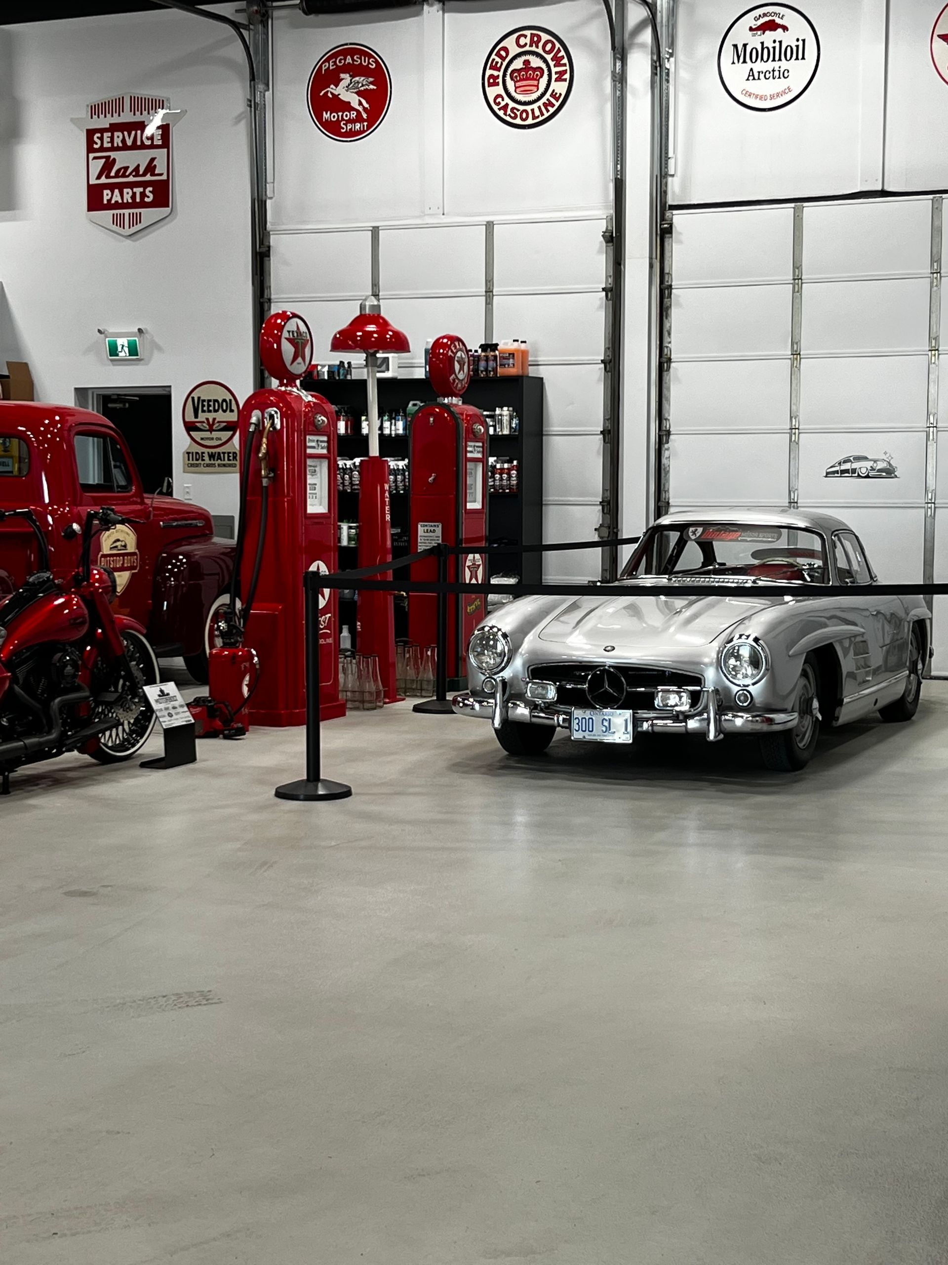 Classic cars in a garage, with a silver car and red gas pumps beside a red vehicle.