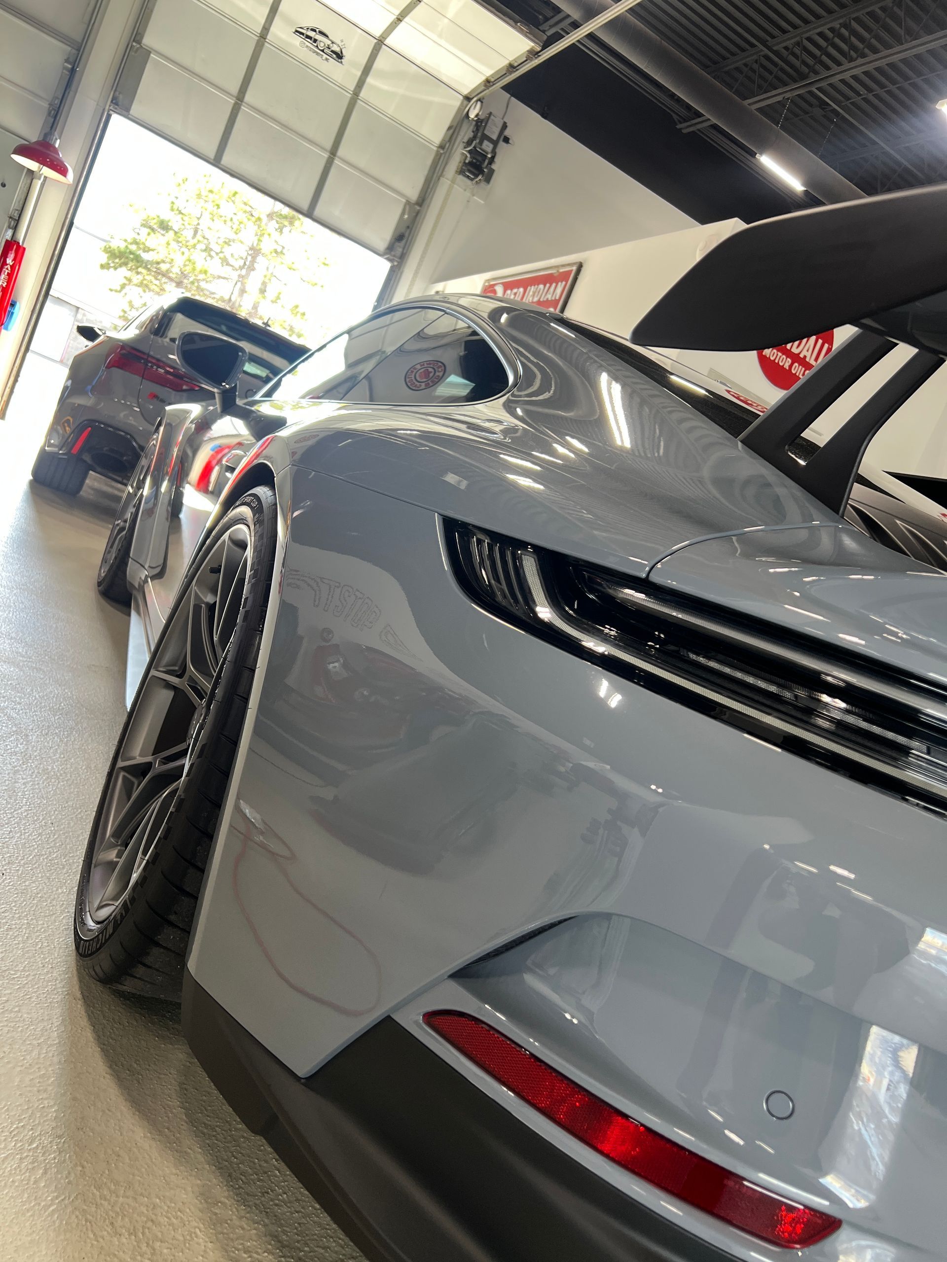 Silver sports car in a showroom, viewed from the rear side with another car parked behind it.