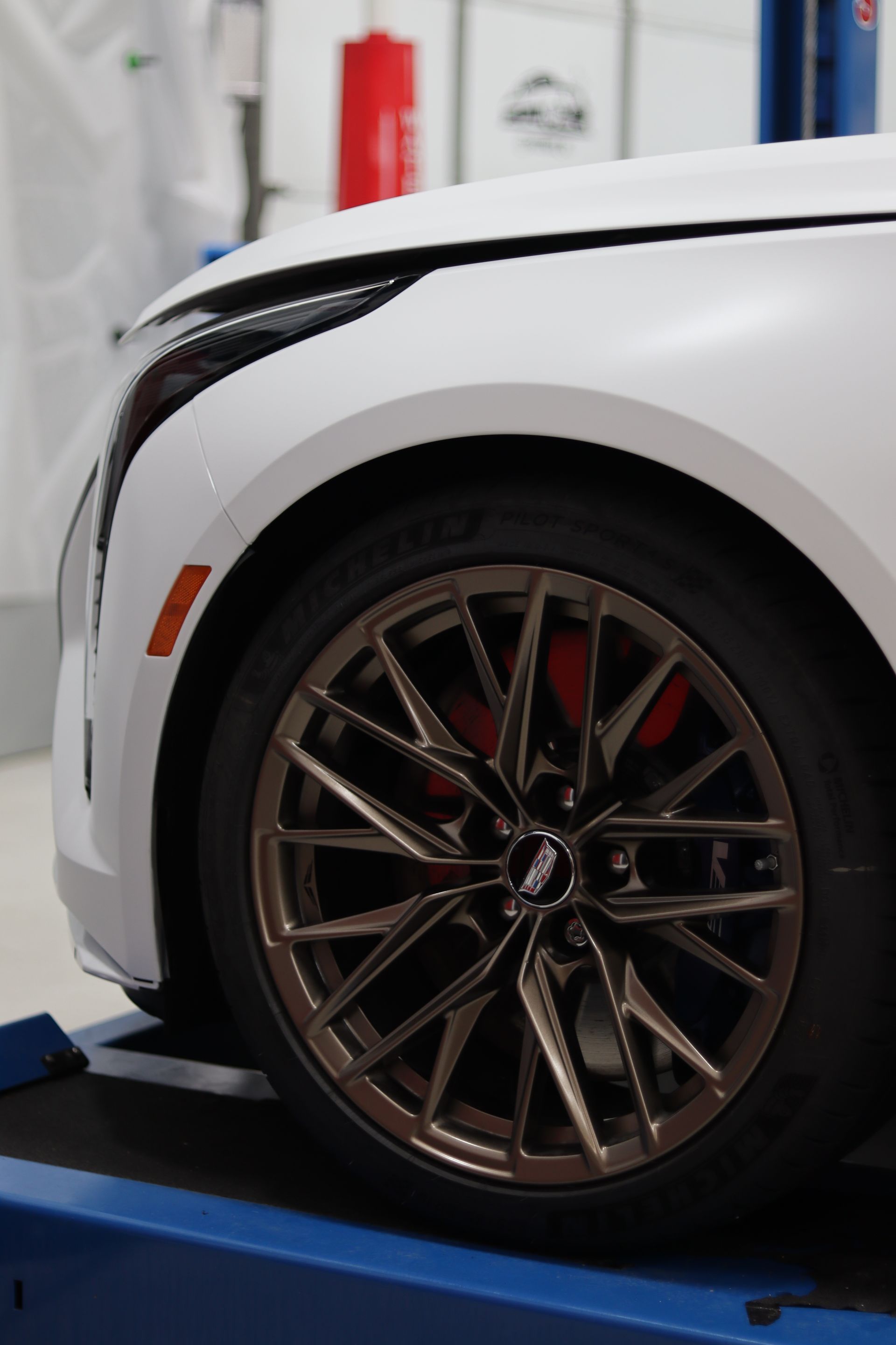 Close-up of a white car’s front wheel on a lift in an auto shop, showing a bronze alloy rim and brake caliper