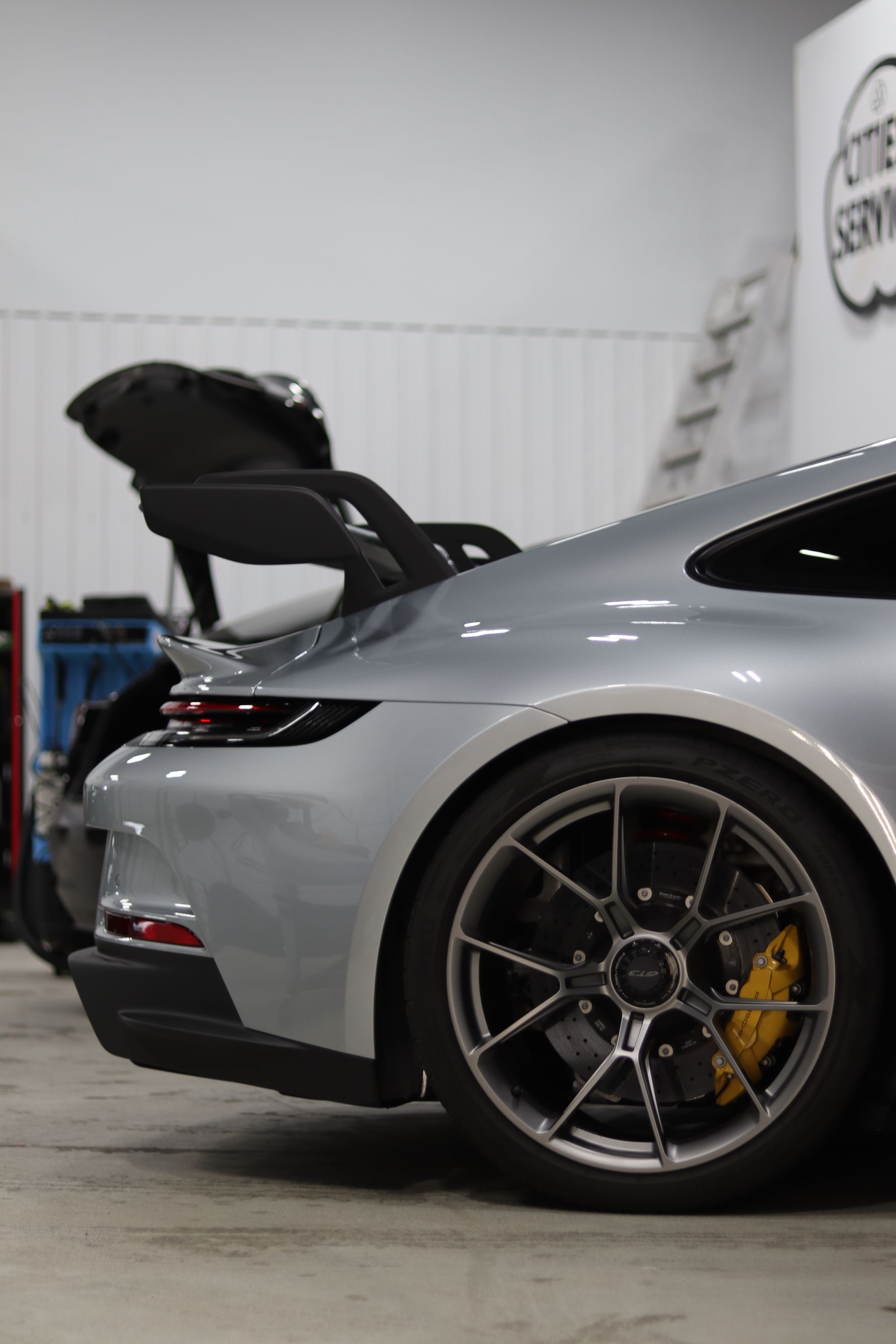Silver sports car with rear hatch open in a garage, showing rear wheel and yellow brake caliper