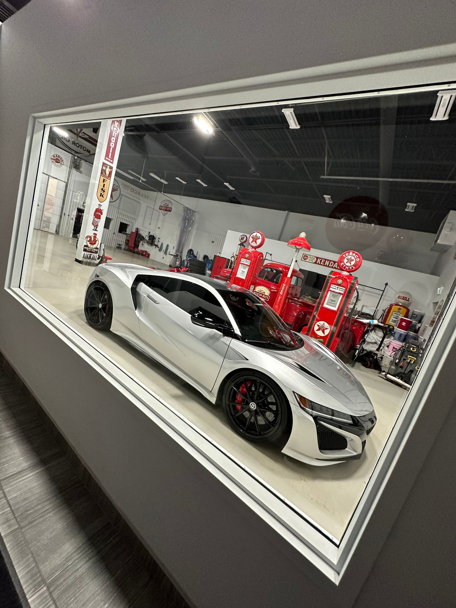 White sports car in a showroom, reflected in a large angled mirror, with red displays in the background.