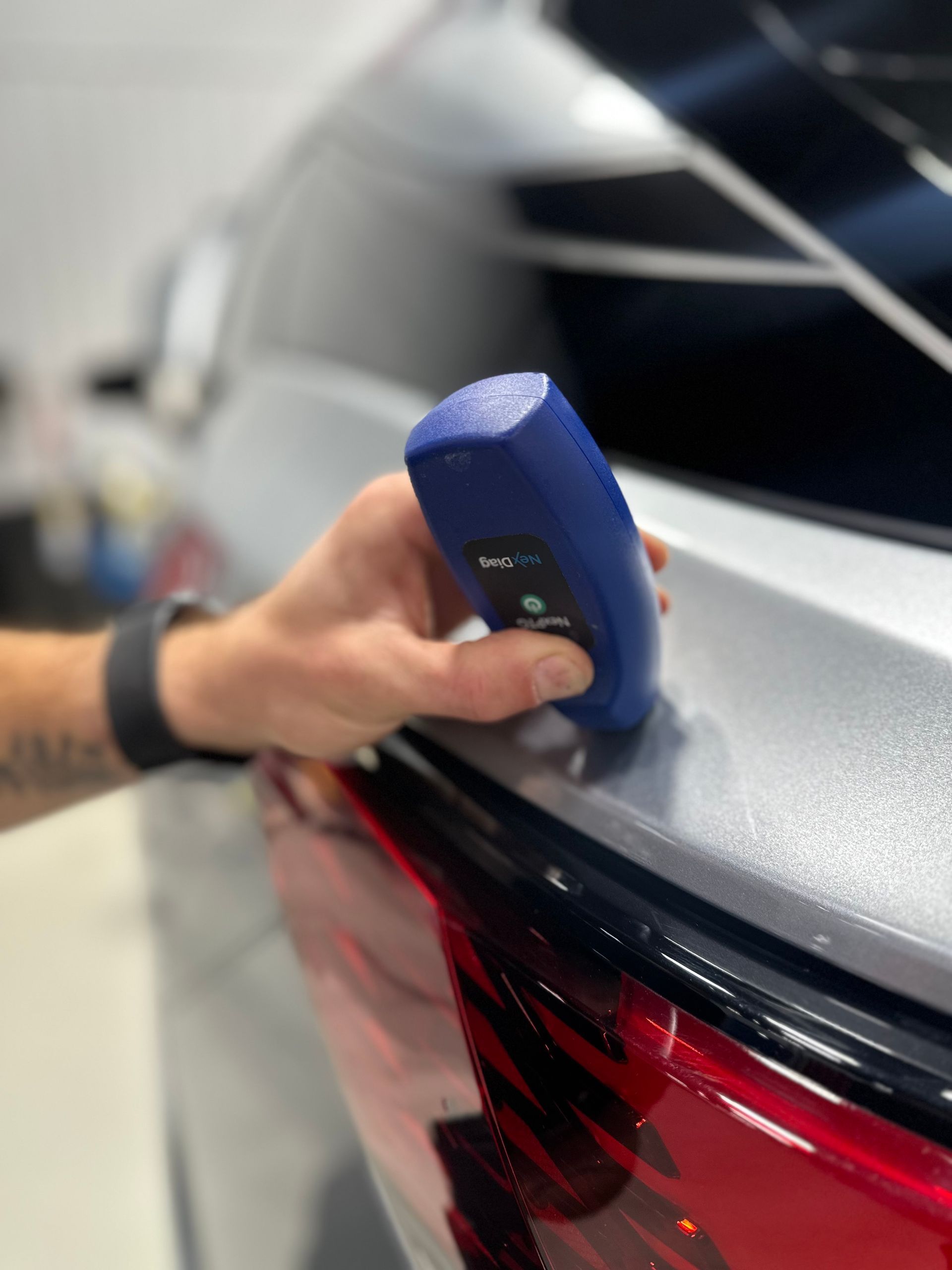 Hand using a blue paint depth gauge on a silver car’s rear panel near the red taillight