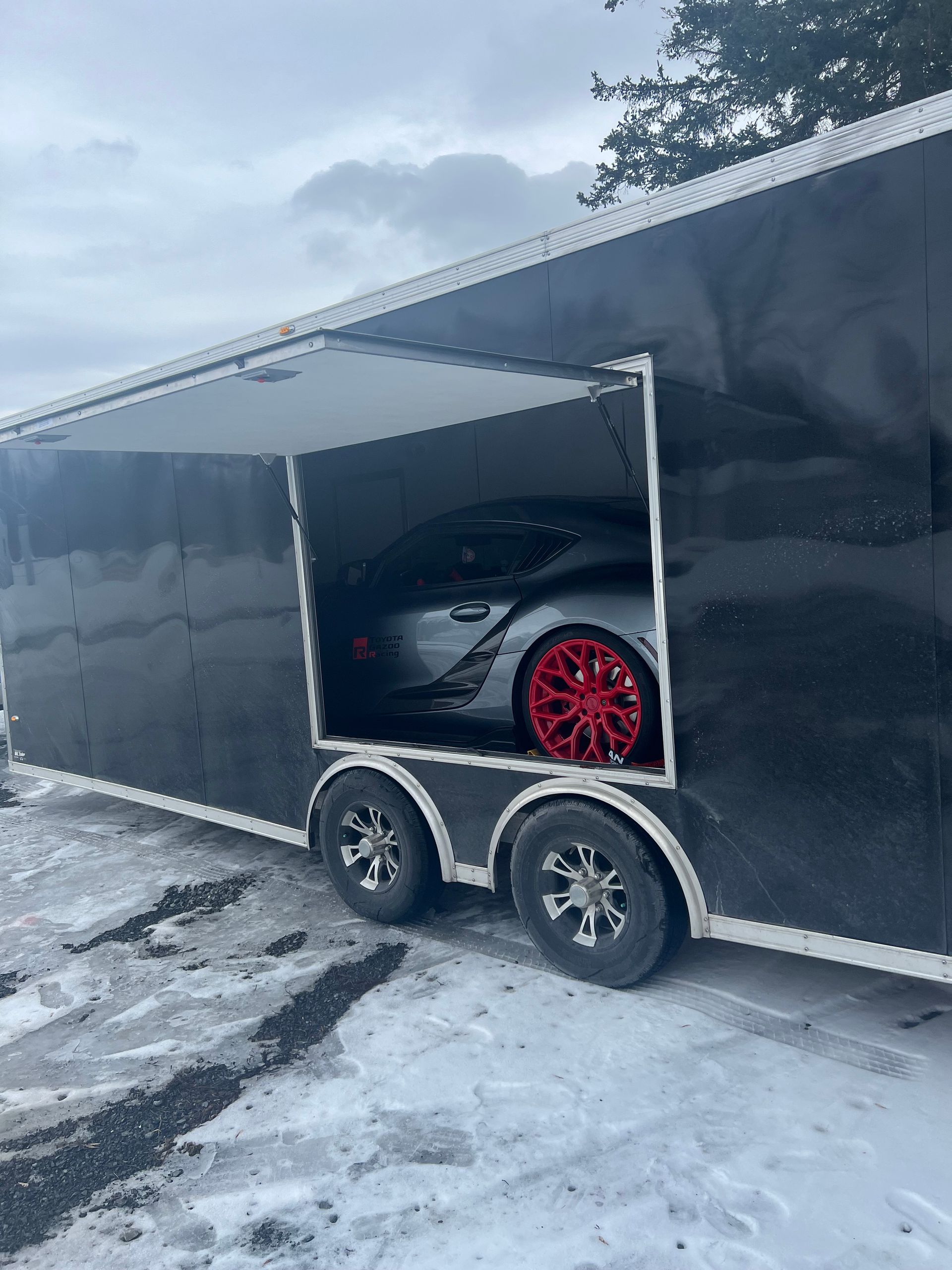 Black sports car with red rims inside a black enclosed trailer on a snowy road.