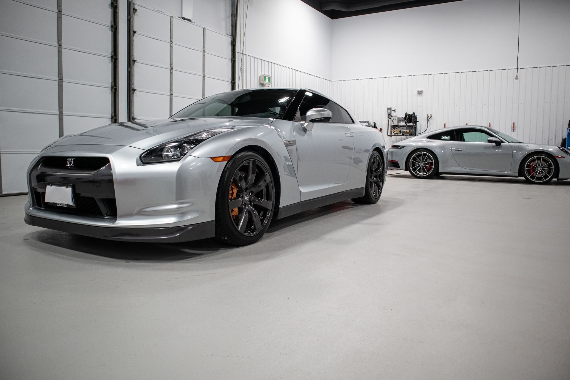 Two silver sports cars in a white garage, one in the foreground and one parked behind.