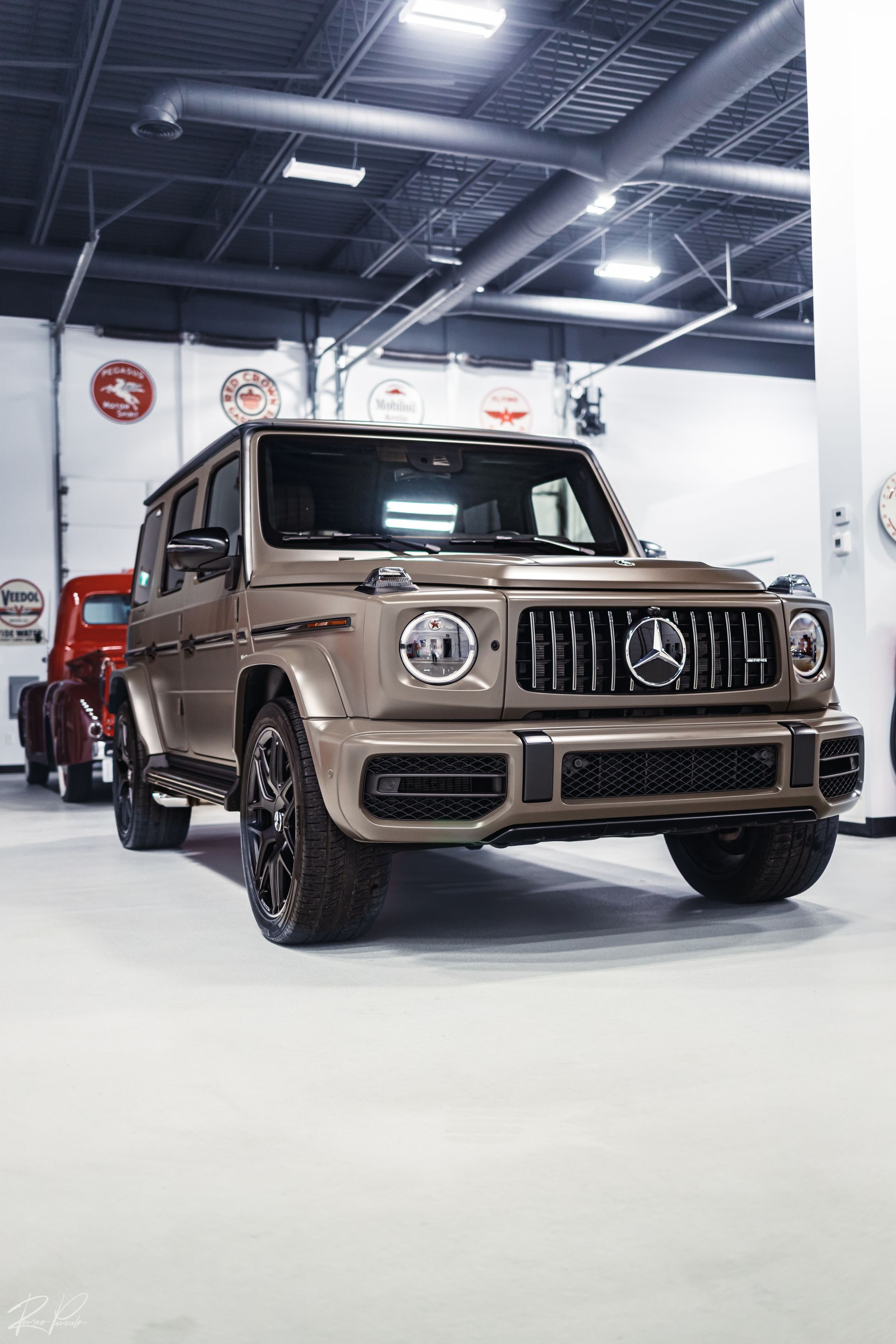 Brown Mercedes G-Class SUV on display in a bright showroom, with a red vehicle behind it.