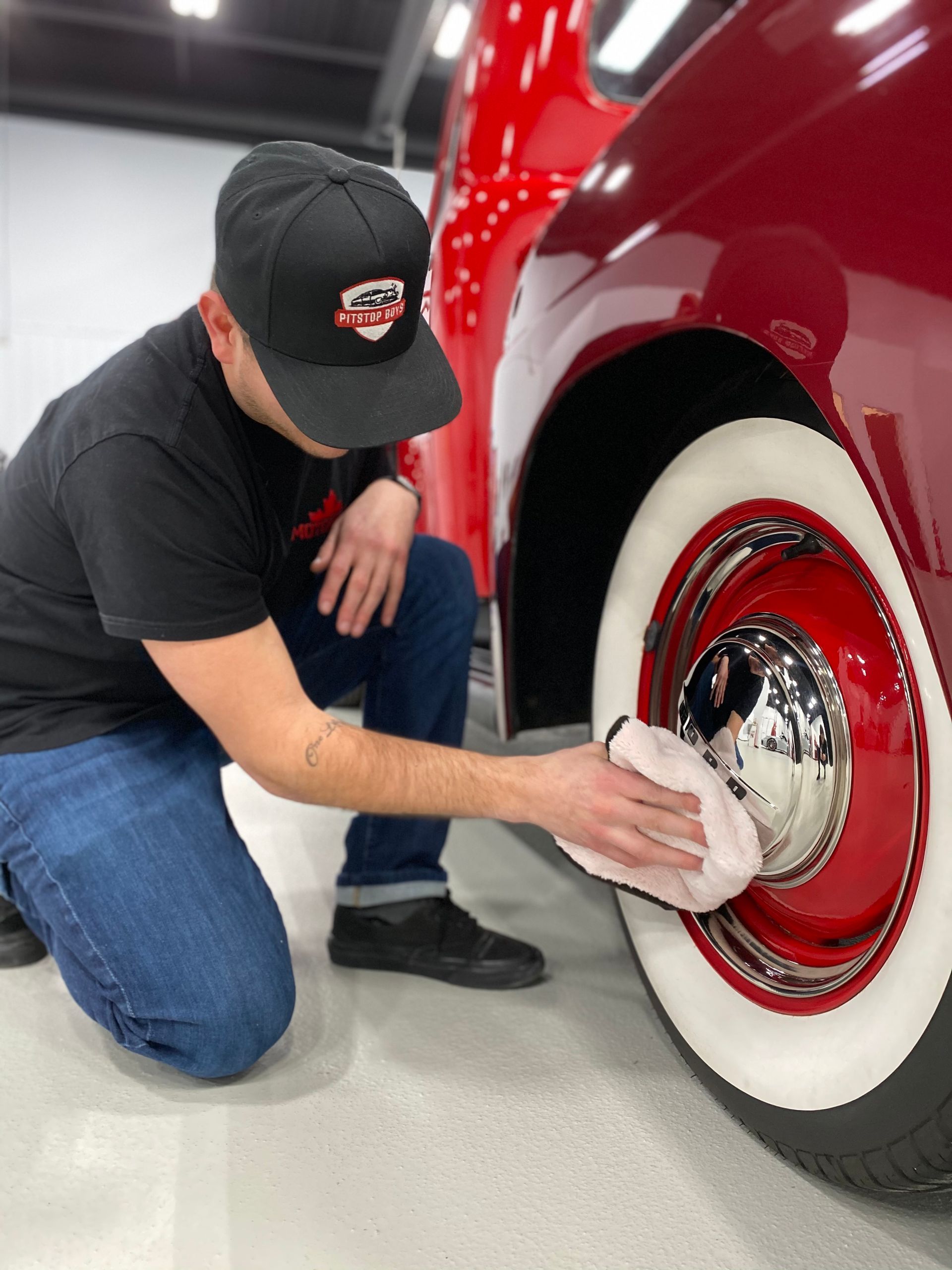 Person kneeling beside a red car, polishing the whitewall tire and chrome hubcap.