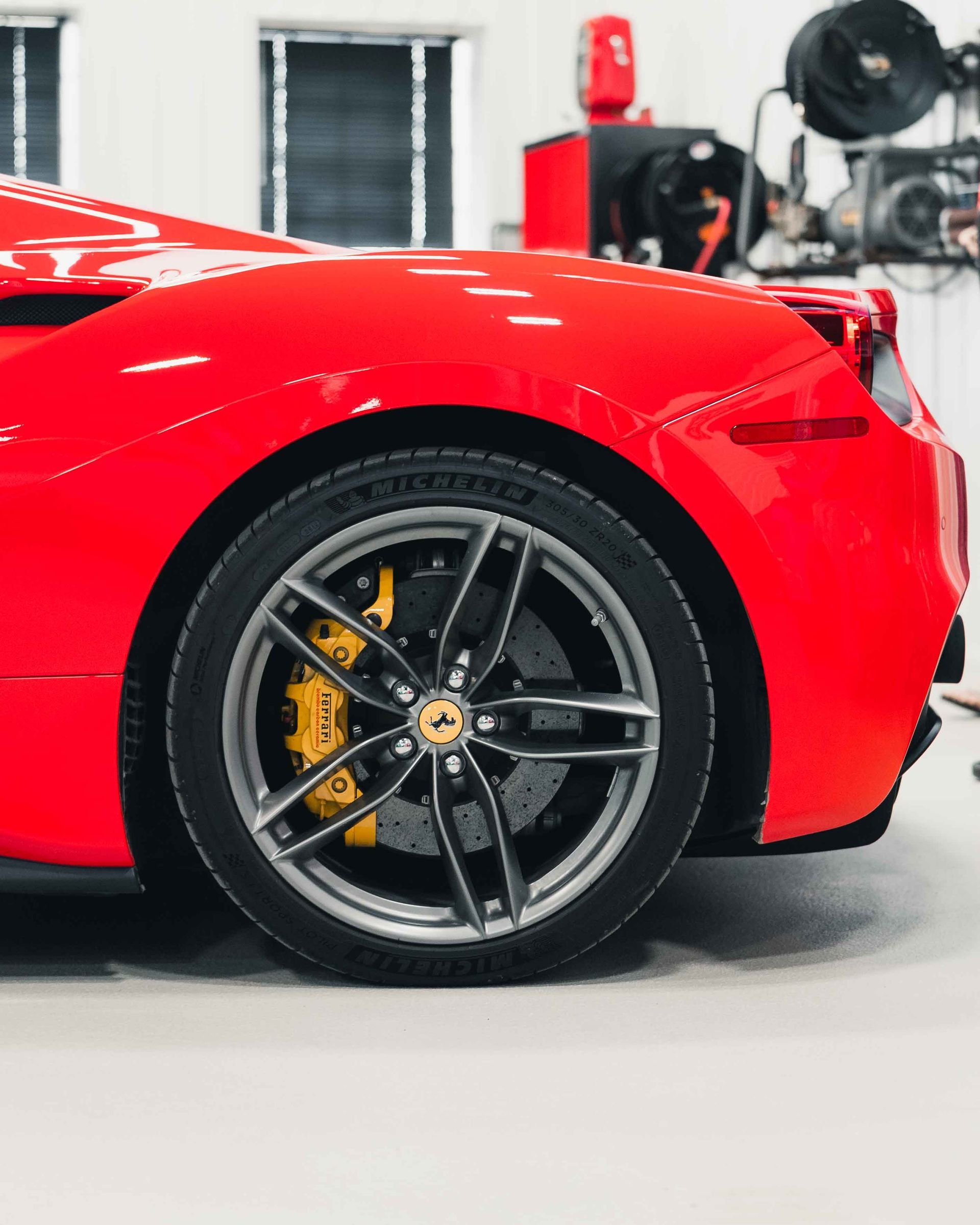 Rear side of a red sports car showing a black wheel with yellow brake caliper in a garage