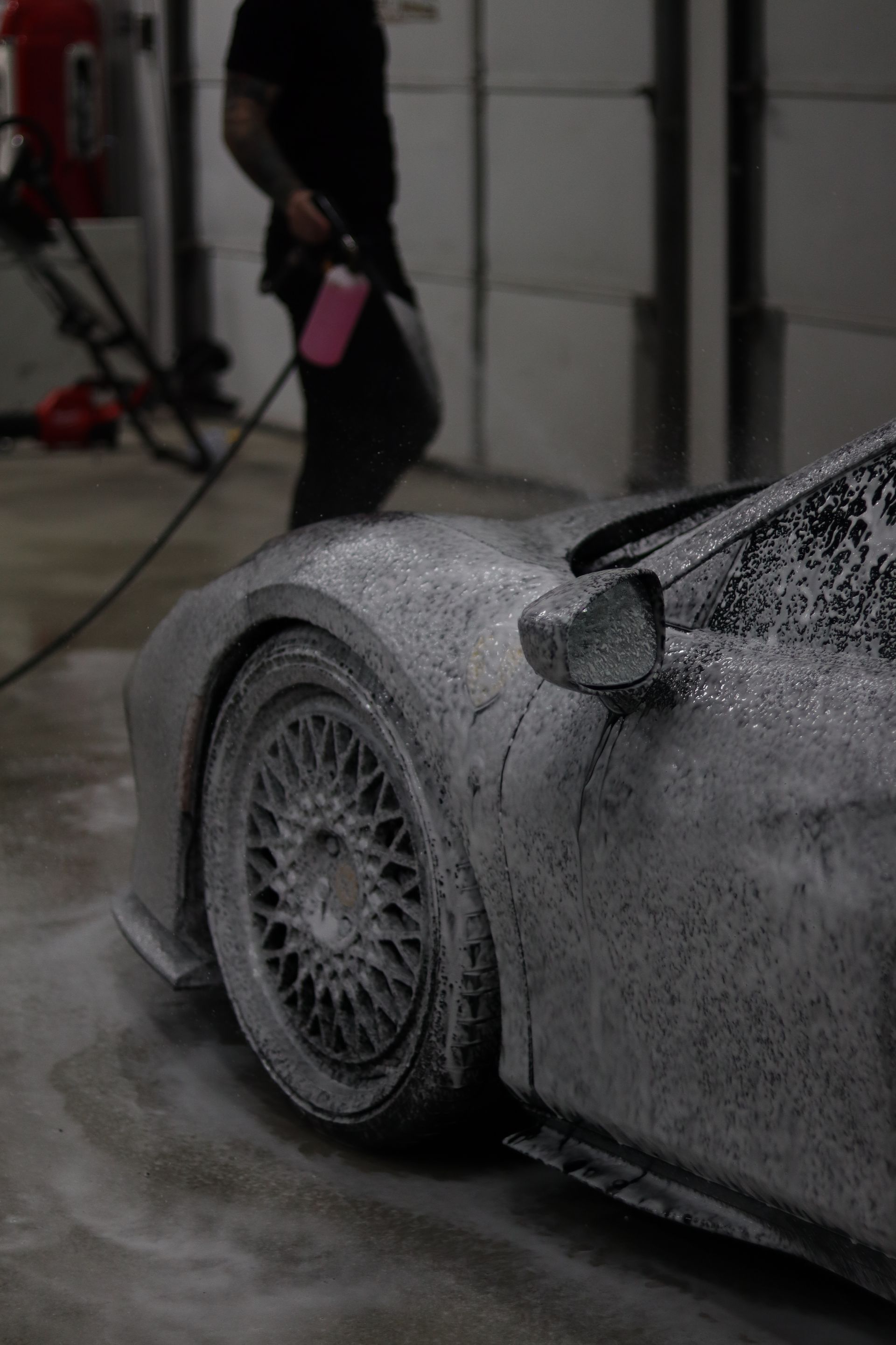Foamy sports car being pressure washed in a garage, with a person spraying it nearby