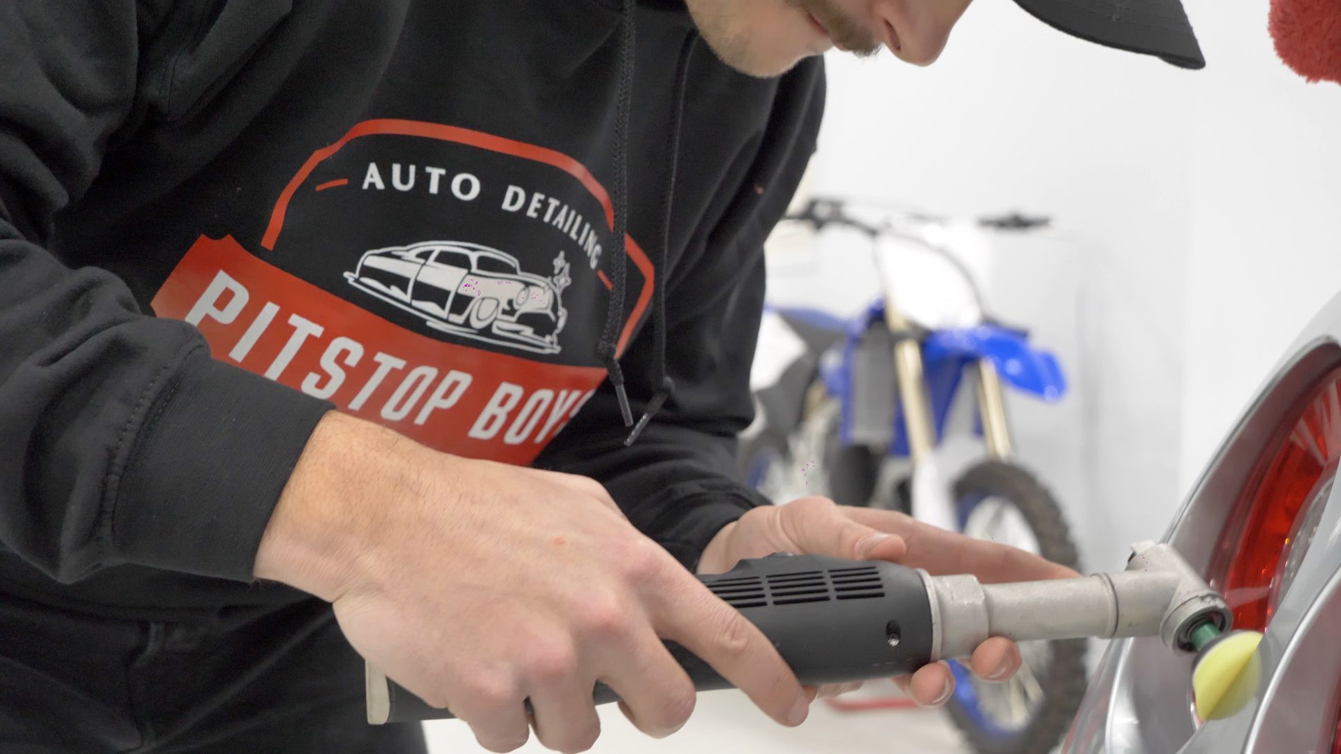 Person using a power drill on a motorcycle in a garage, wearing a black Pitstop Boy shirt.
