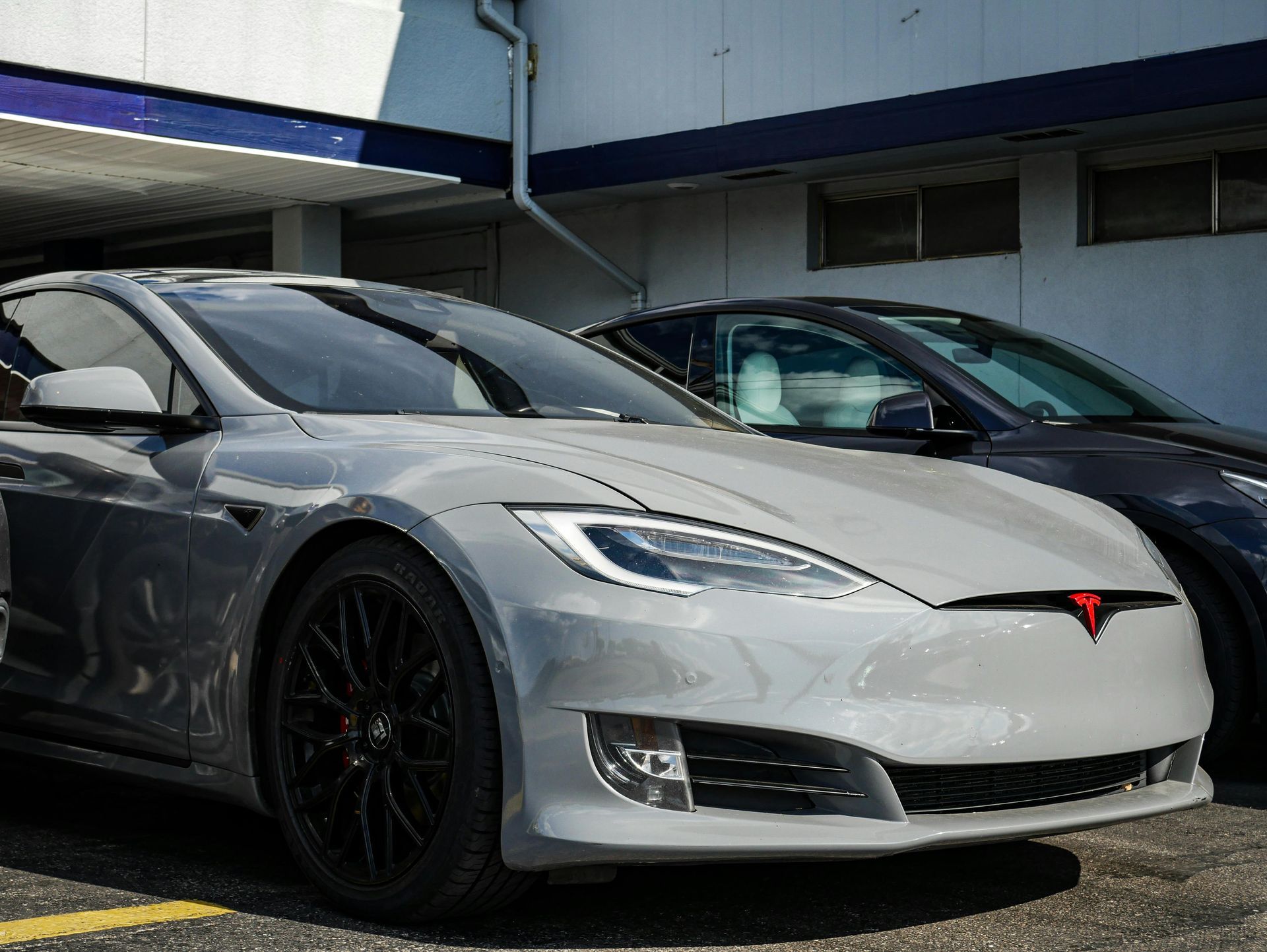 Silver Tesla sedan parked outside a building with a black front wheel and red logo.