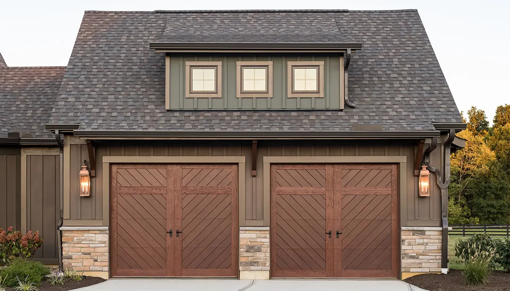 Two-car garage with brown wooden doors, stone accents, and dormer windows on a gray house façade