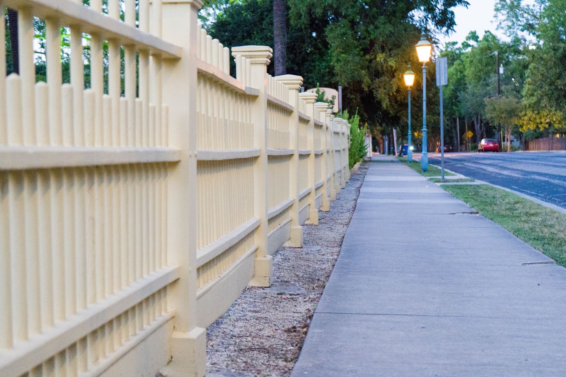 Cream-colored picket fence borders a sidewalk, street, and trees. Evening lighting illuminates the scene.