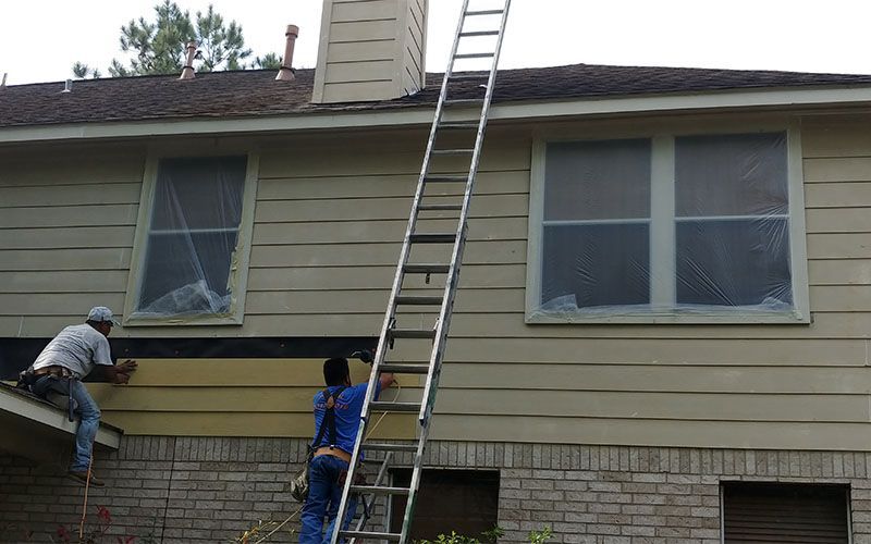 Man in a blue cap, hammering siding onto a building.