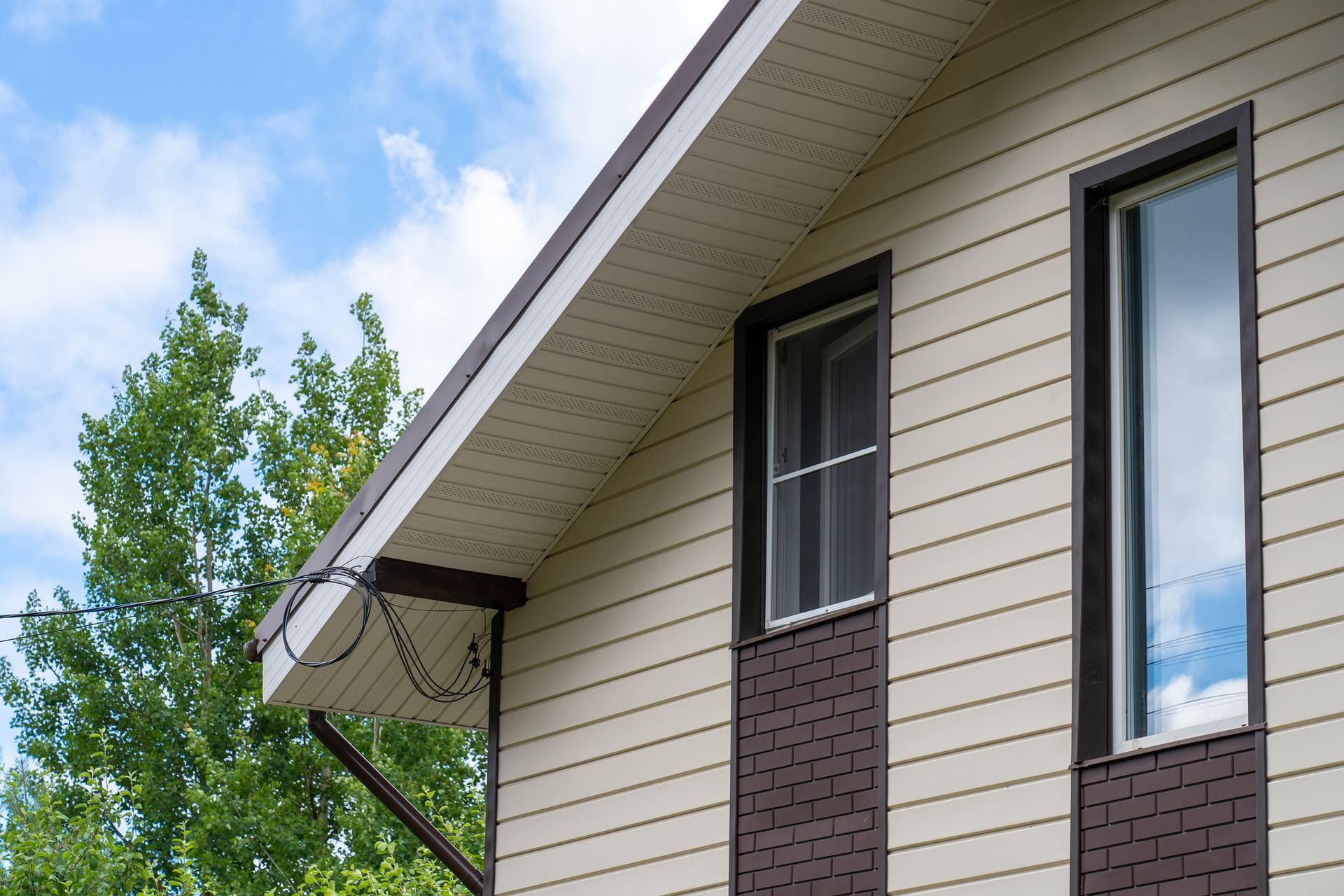 Tan and brown house siding with two windows and a roof against a cloudy sky.