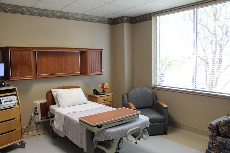 A hospital room with a bed , chair , desk and window.