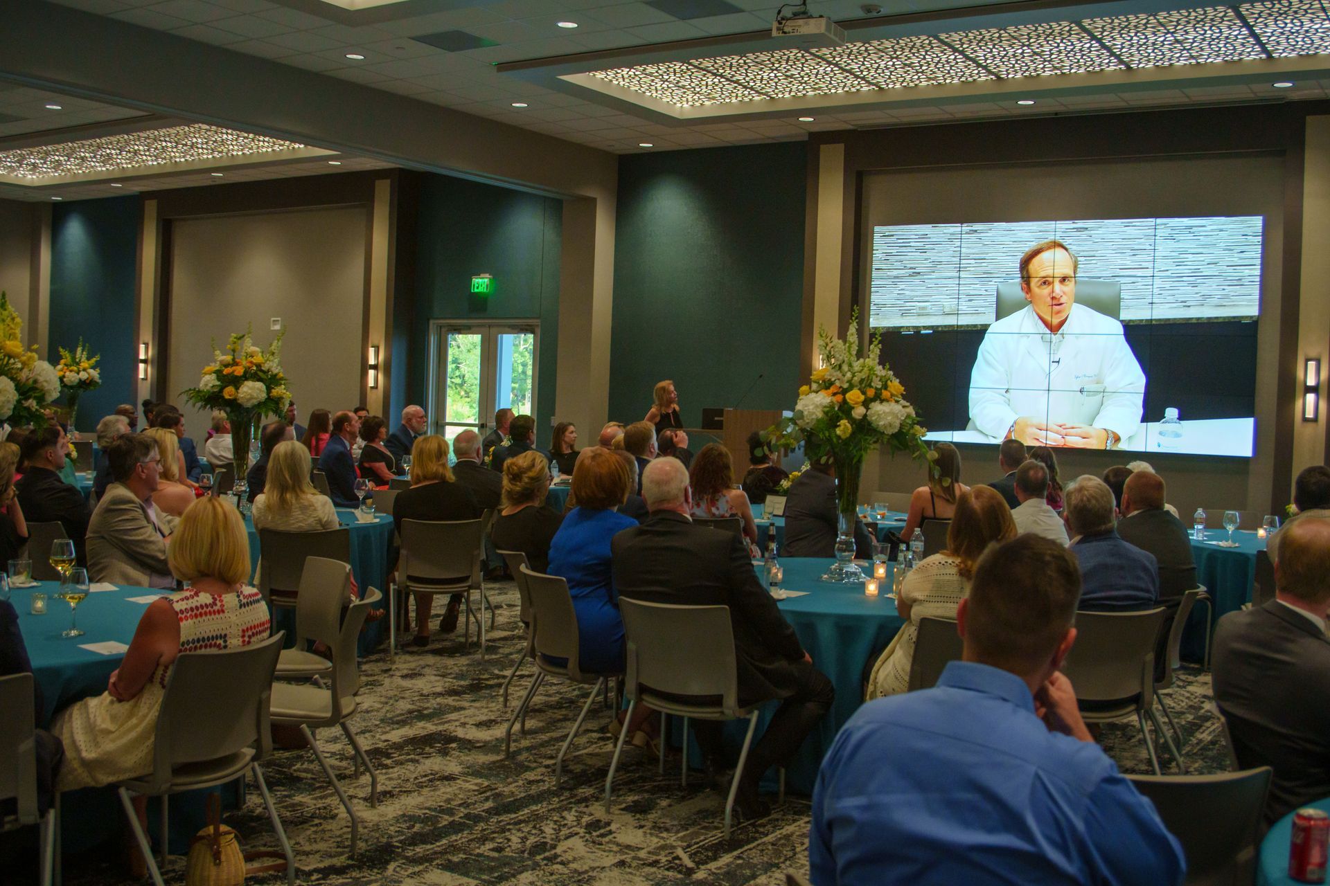 A group of people are sitting at tables in a large room watching a presentation on a large screen.