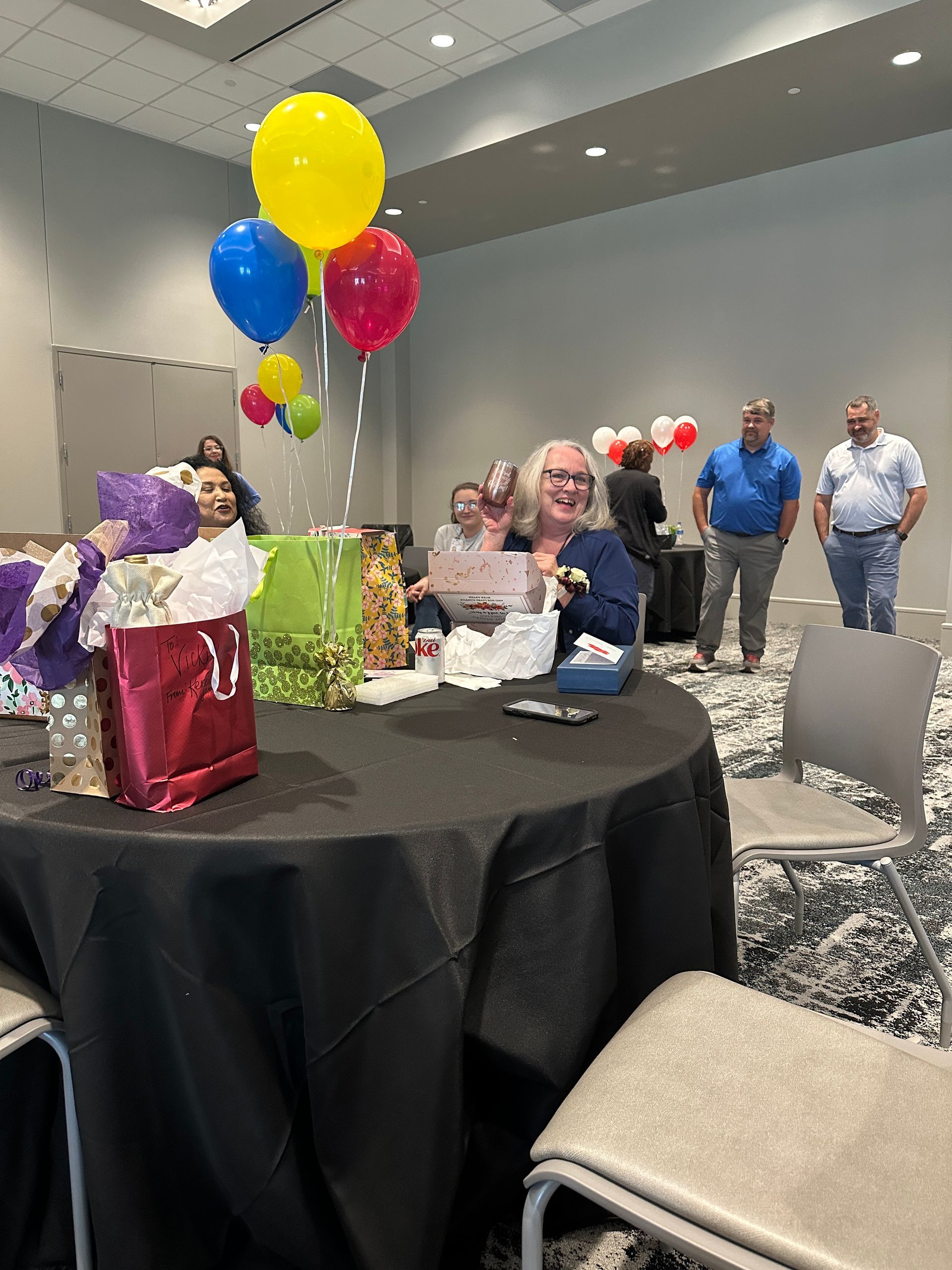 A group of people are sitting around a table with balloons and presents on it.