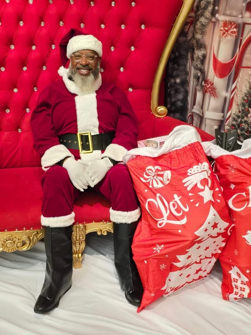 man dressed as santa claus sitting in an oversized red chair with a bag of gifts next to him