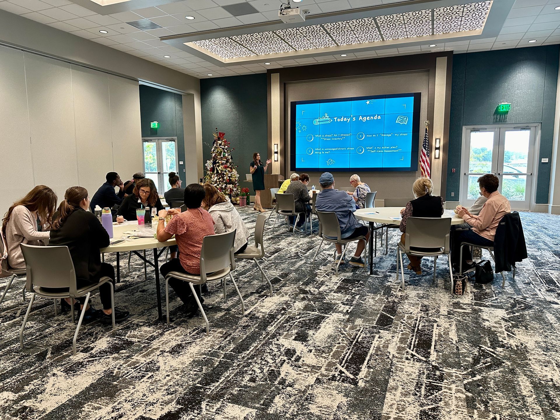 A group of people are sitting at tables in a large room with a christmas tree in the background.