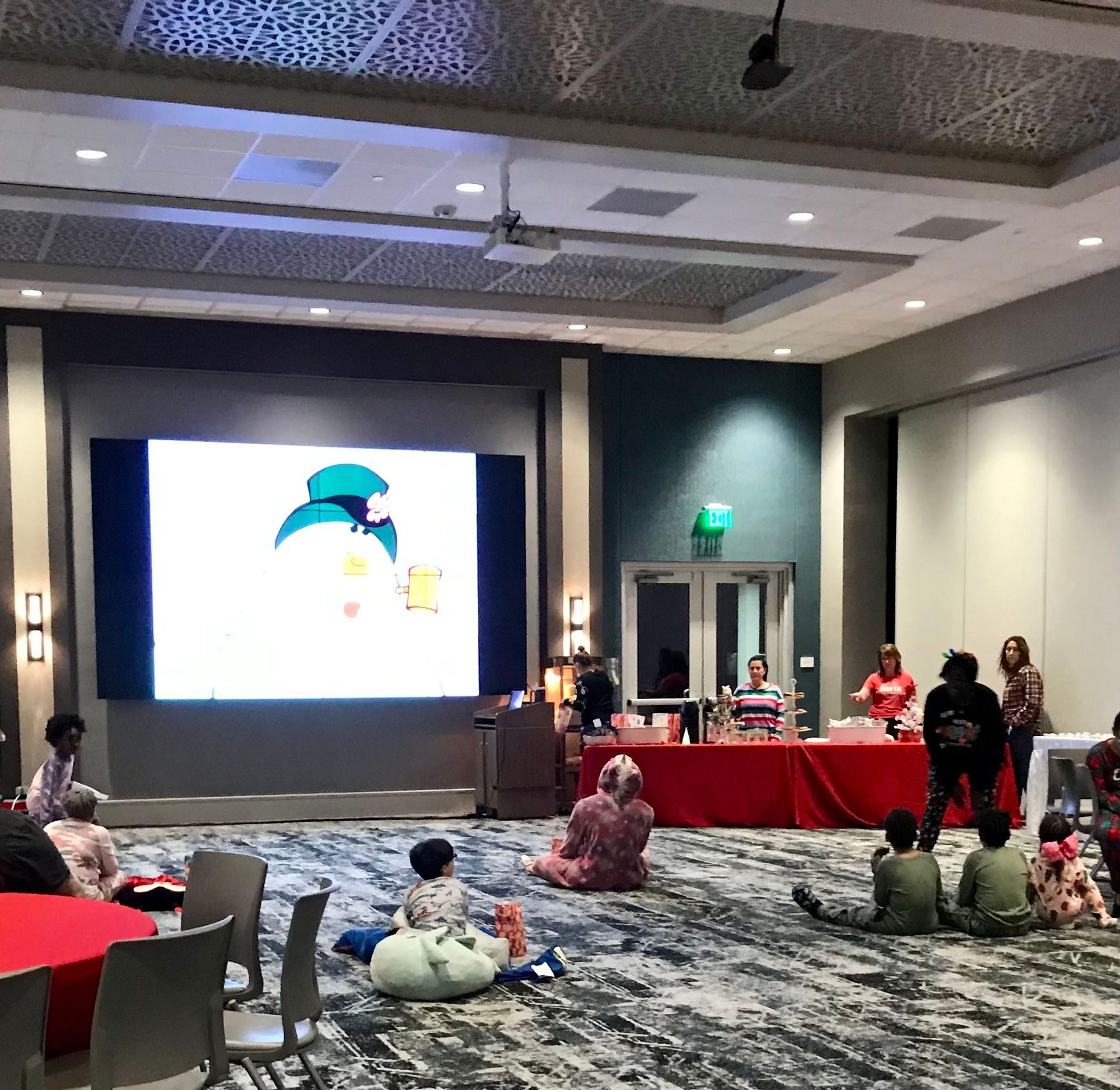 A group of children are sitting on the floor in front of a large screen with a snowman on it
