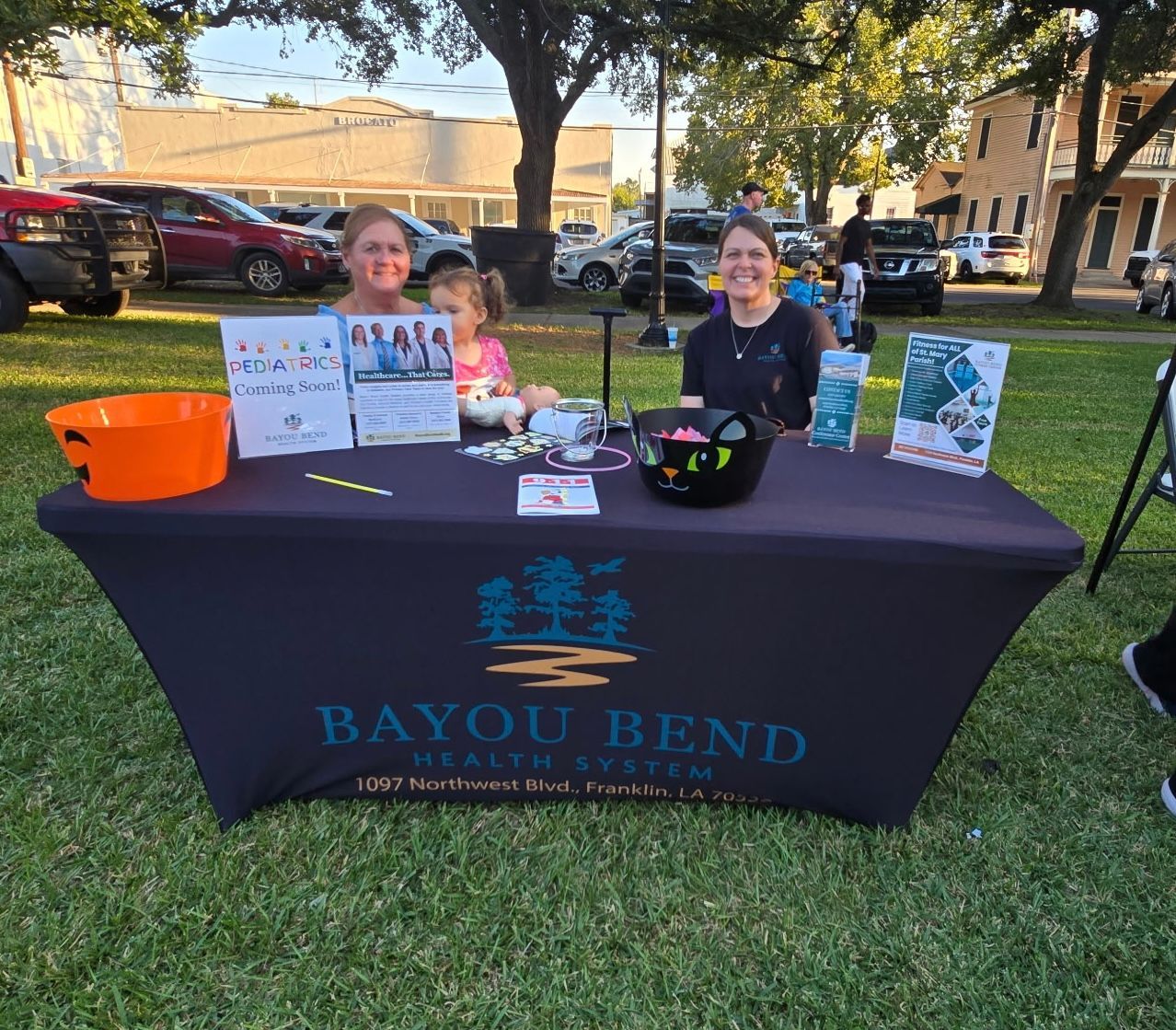 2 women and a child sitting at an information table outside at a park