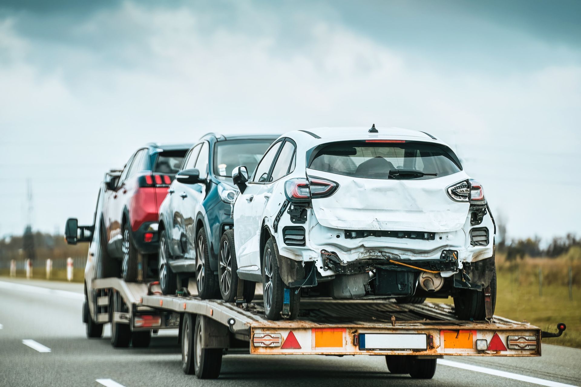 Cars loaded on a car carrier trailer, including a damaged white hatchback, traveling on a highway.