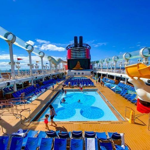 Cruise ship pool deck with swimmers, blue lounge chairs, and a giant screen under a bright sky