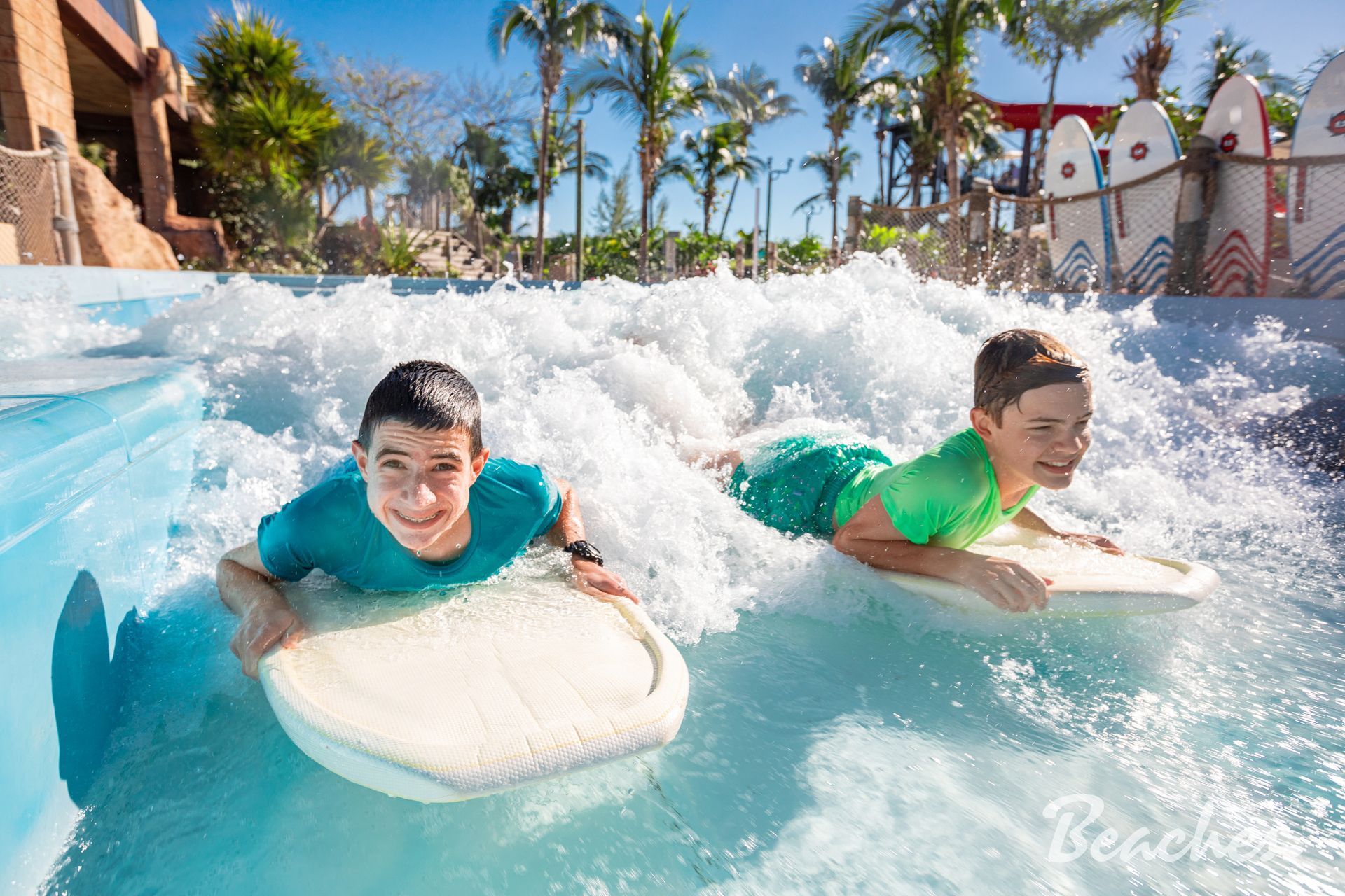 Two people bodyboarding through blue water at a sunny water park, smiling in the surf
