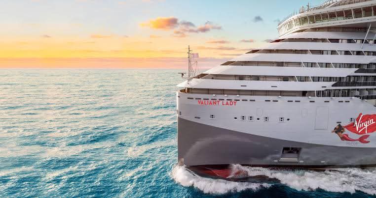 Large white cruise ship sailing on turquoise water at sunset, with a colorful sky and waves at the bow