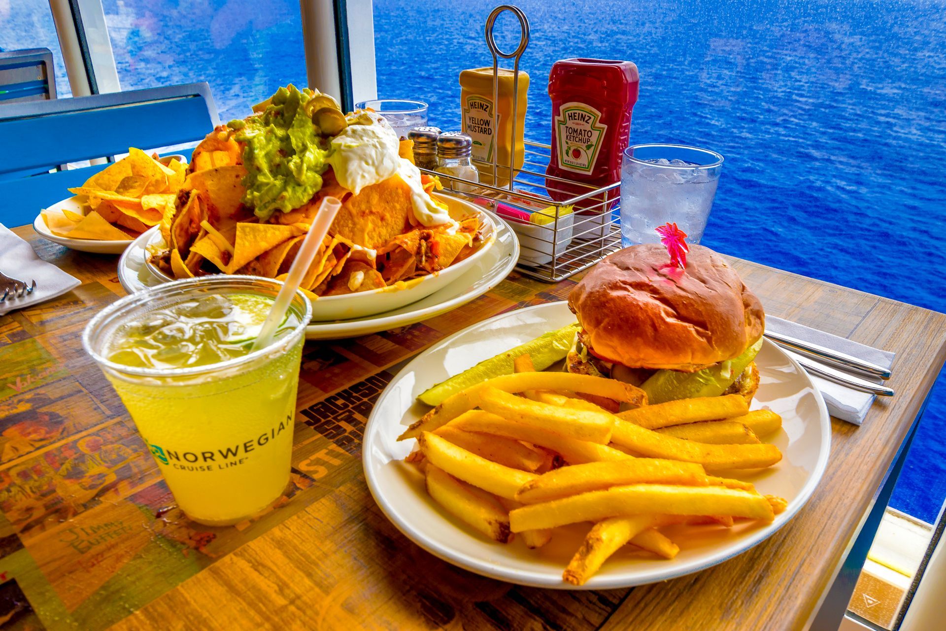 Two plates of fries and burgers with a drink on a wooden table by a blue waterfront.