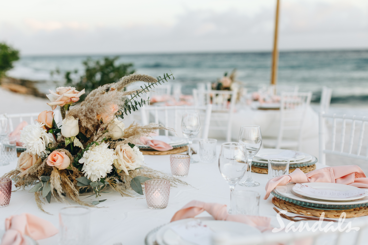 Elegant beachside table with pastel floral centerpiece, glassware, and plates set for an outdoor seaside dinner