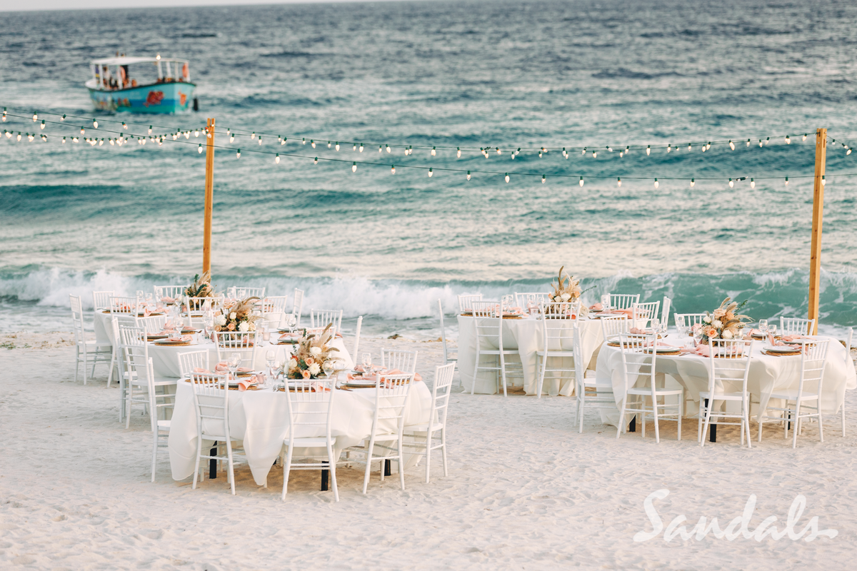 Beach wedding reception with white tables and chairs by the ocean, with waves and a boat in the background