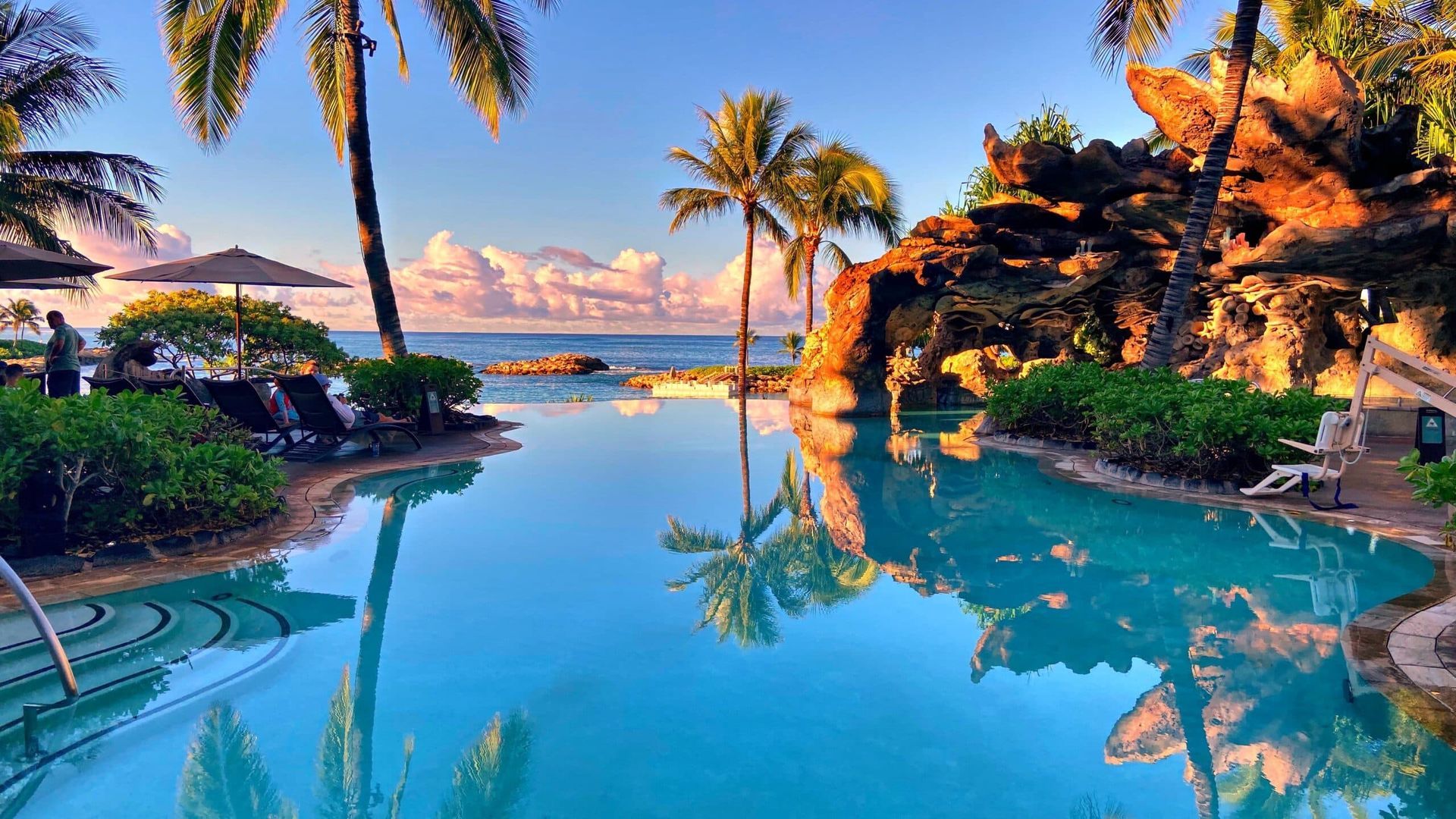 Tropical resort pool with palm trees, rock waterfall, and ocean sunset reflections