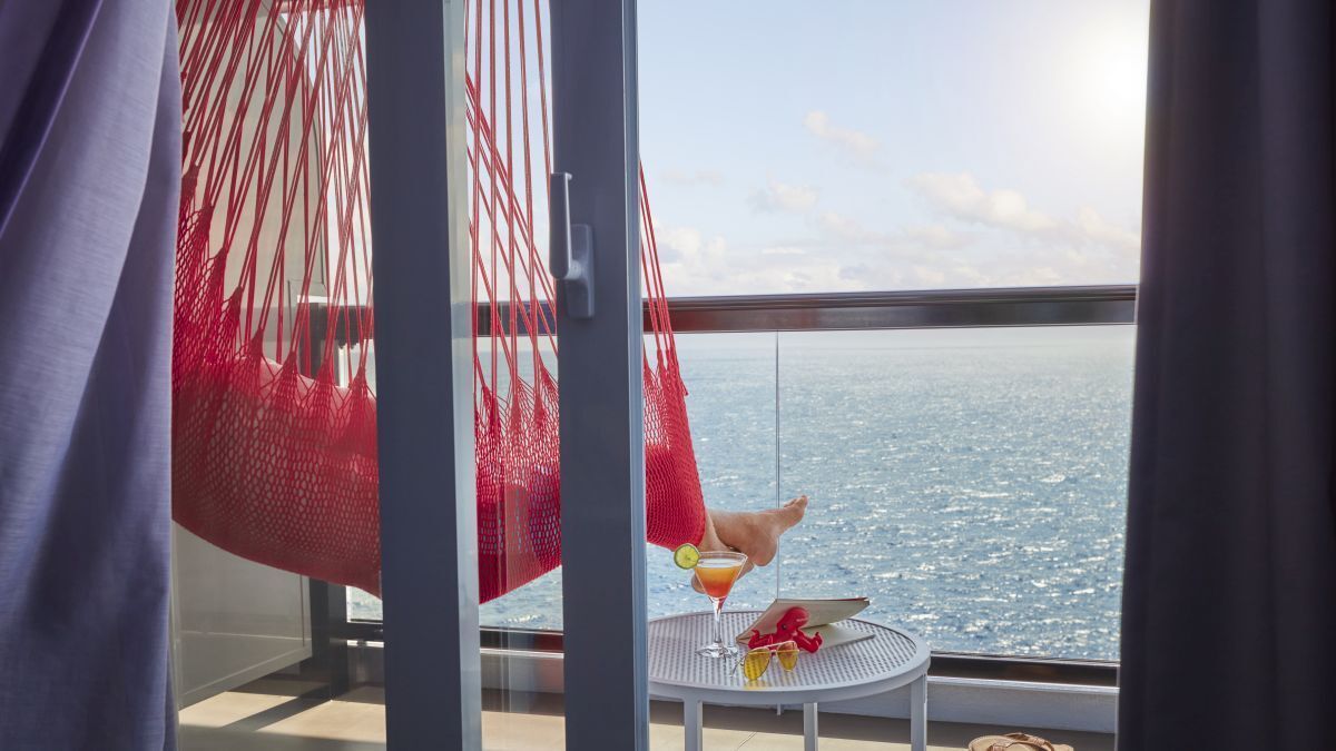 Balcony table with fruit and drink overlooking a calm ocean at sunset, framed by curtains and a red hammock