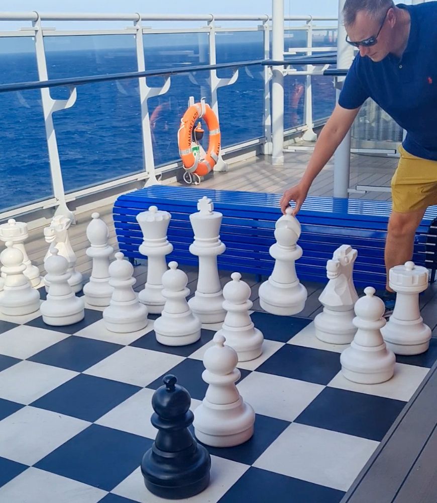 Person moves a white chess piece on a giant outdoor chessboard aboard a ship.