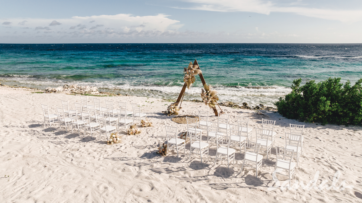 Beach wedding setup with white chairs, wooden arch, turquoise ocean, and green shrub under bright sun