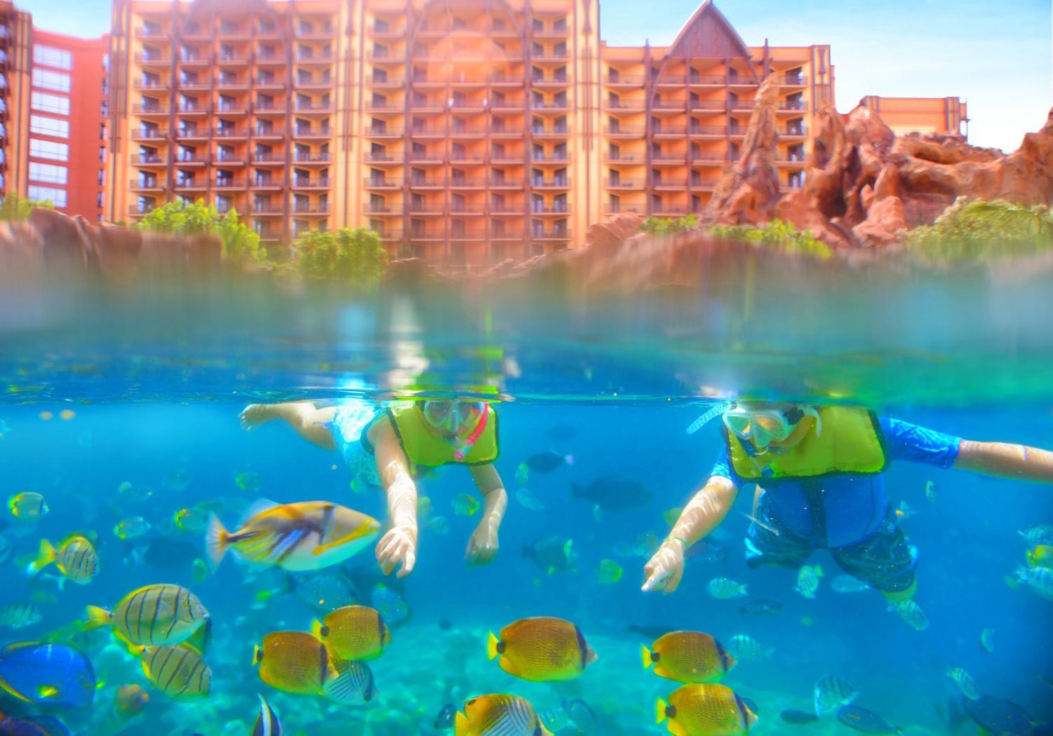 Snorkelers swimming above colorful fish in a turquoise lagoon, with a resort hotel behind them