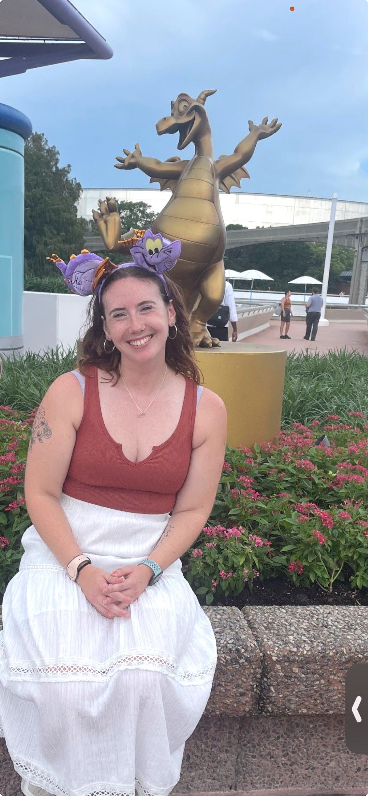 Smiling woman in red top and white skirt seated before a dragon statue in a park
