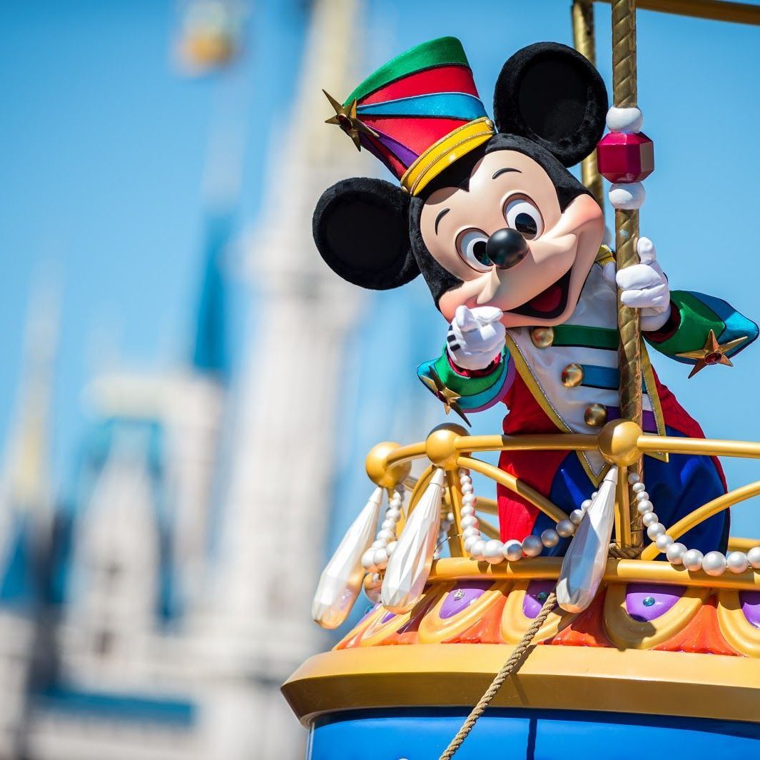 Mickey Mouse in a colorful parade outfit waving from a decorated float against a blue sky