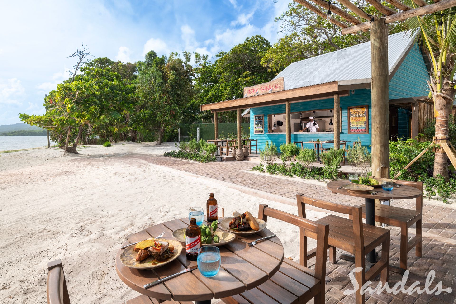 Beachfront dining area with wooden table, tropical trees, and a blue-roofed hut by the shore