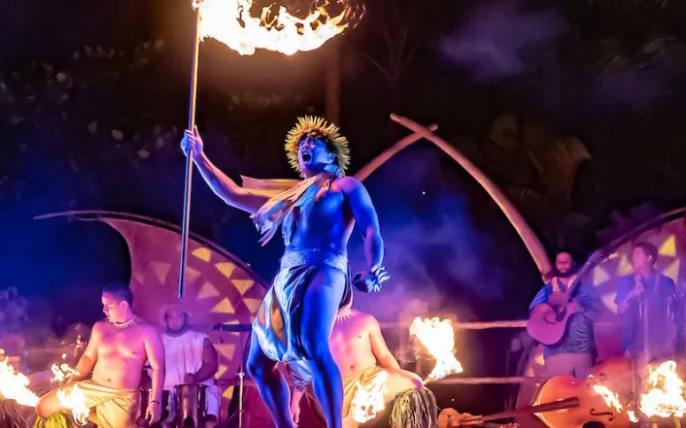 Fire performer on a stage with bright flames and purple lighting, holding a flaming staff.