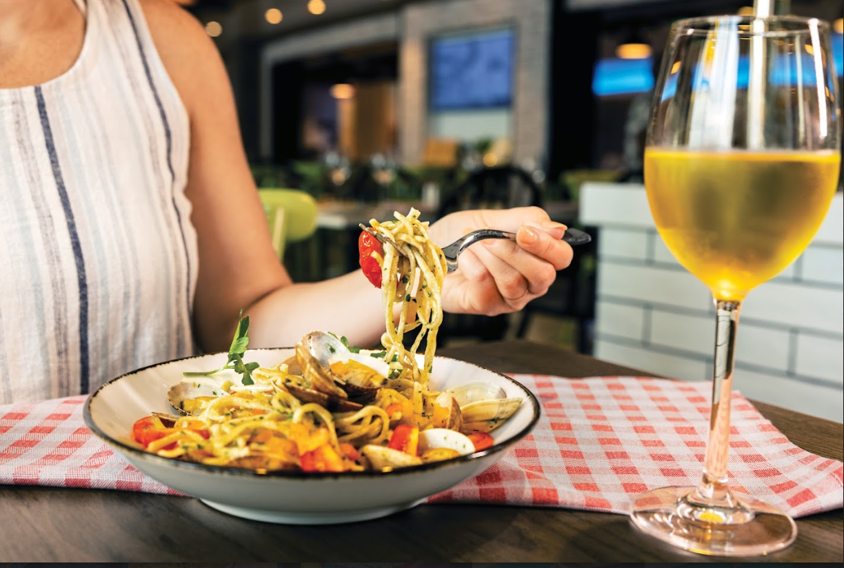Person eating pasta at a restaurant table with a glass of white wine